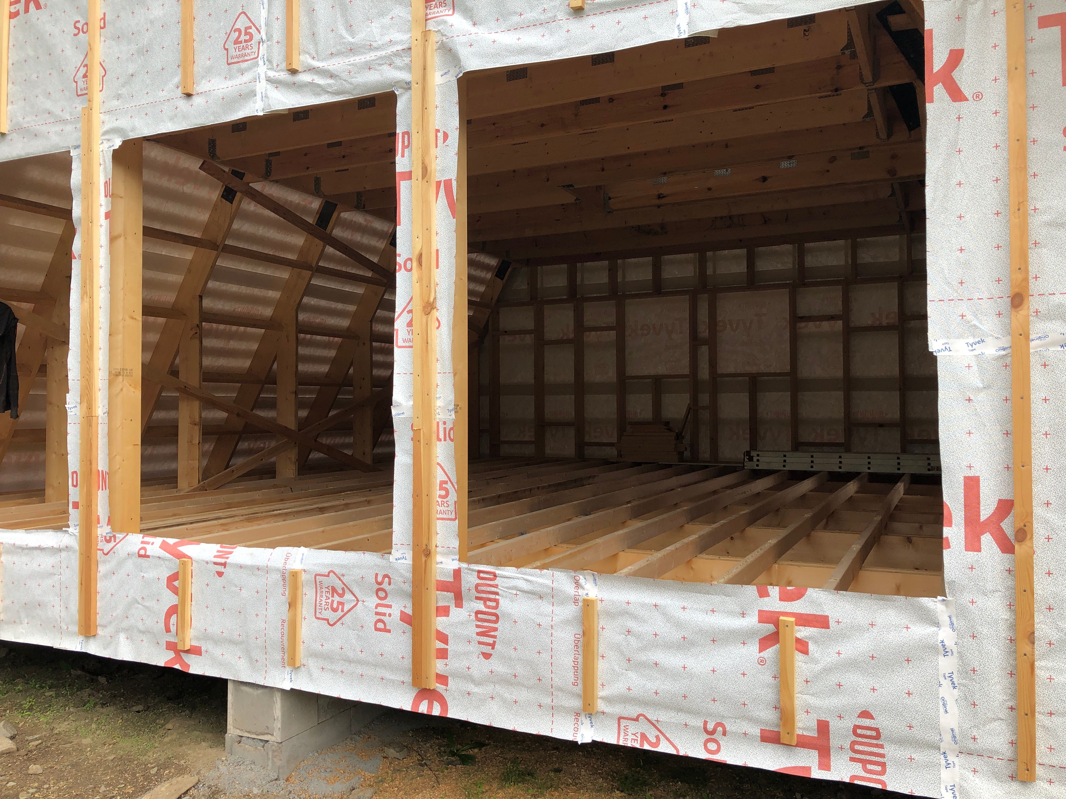 An exterior close-up view of an Avrame kit home during the structural phase of a house construction project. The image highlights the raw wood floor joists, ceiling beams, and sloped wall studs exposed through large window openings. The exterior structure is wrapped in protective white DuPont Tyvek weather barrier paper with red lettering, sitting elevated on concrete block foundations.
