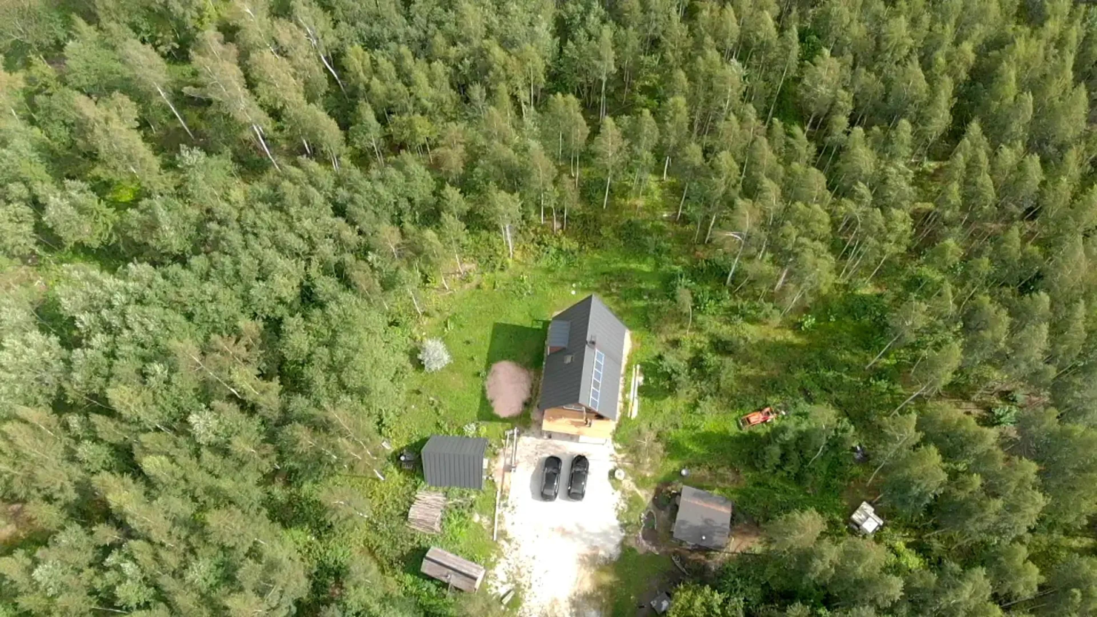 An aerial drone shot shows a dark-roofed A-frame house with solar panels nestled in a lush forest clearing alongside several small outbuildings and two parked cars.