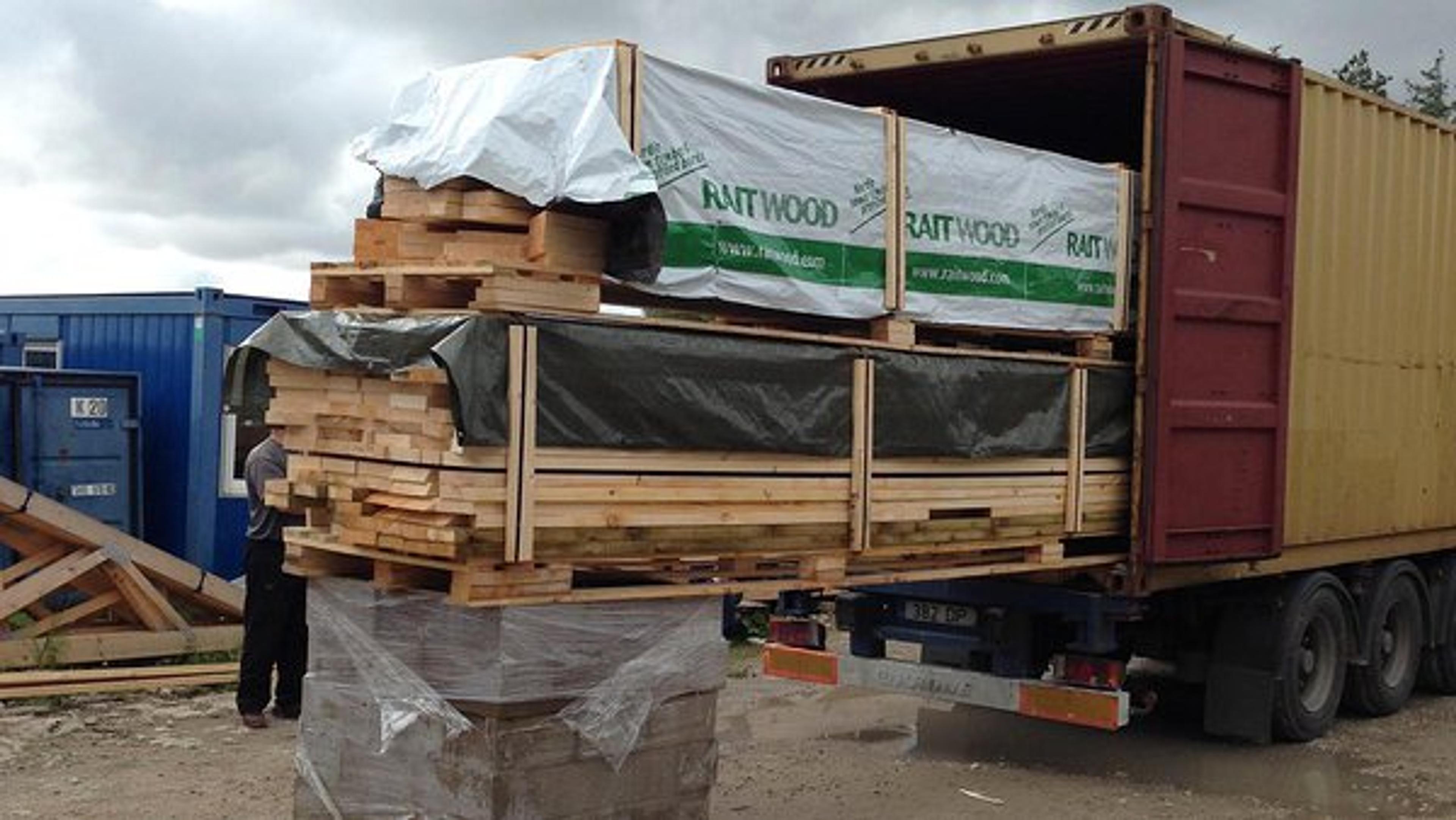 Large bundles of pre-cut lumber labeled "Raitwood" are being loaded into a yellow shipping container on a flatbed trailer.