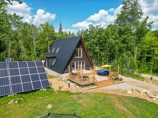 An aerial view shows a modern black A-frame cabin powered by a large ground-mounted solar array, featuring an expansive wooden deck with lounge seating nestled in a lush green forest.