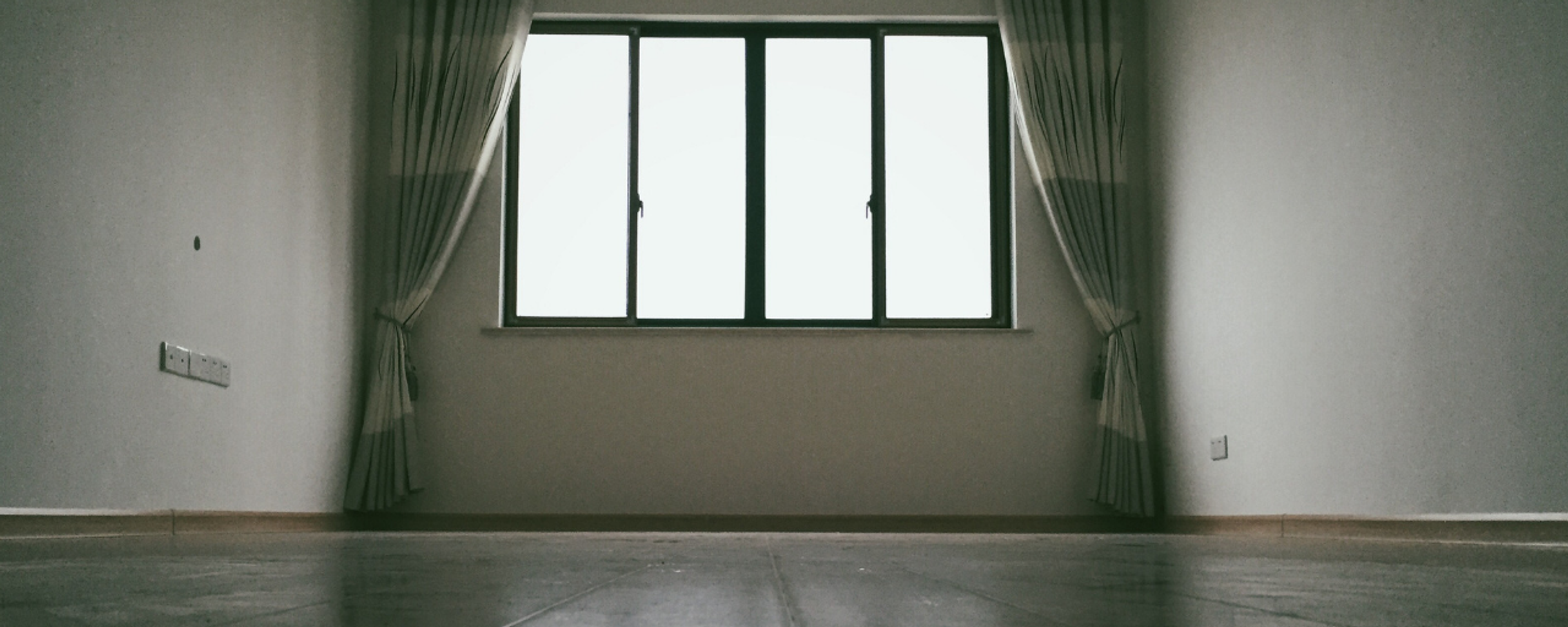 An empty, dimly lit room features a large central window with drawn-back curtains, casting a bright glow onto the wooden floor.