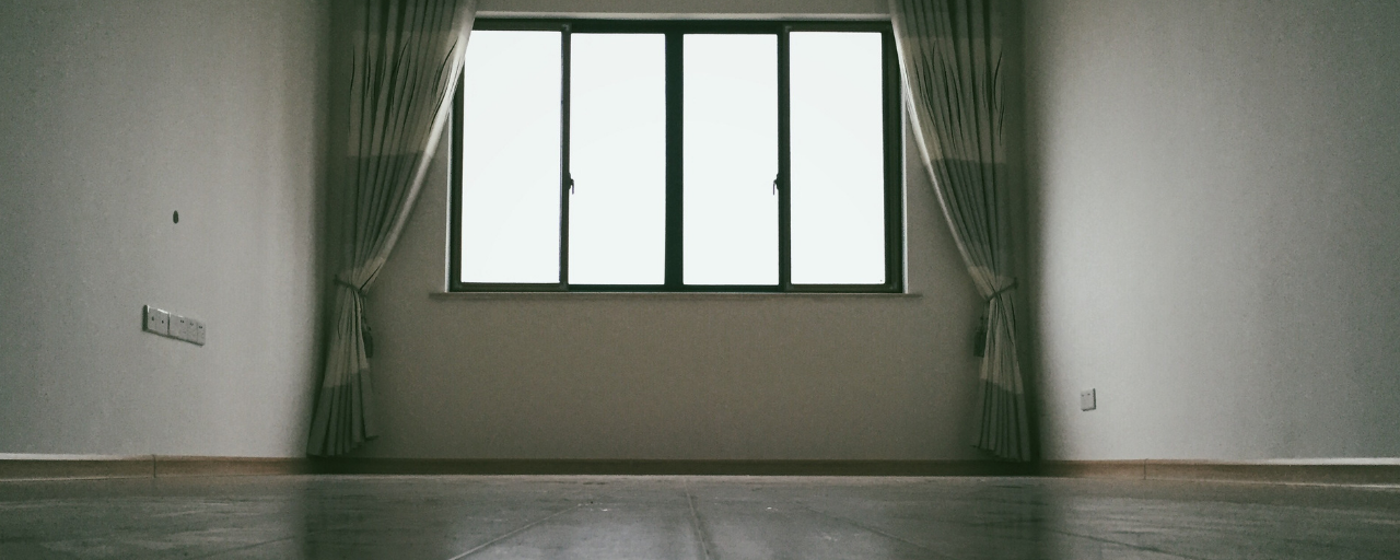 An empty, dimly lit room features a large central window with drawn-back curtains, casting a bright glow onto the wooden floor.