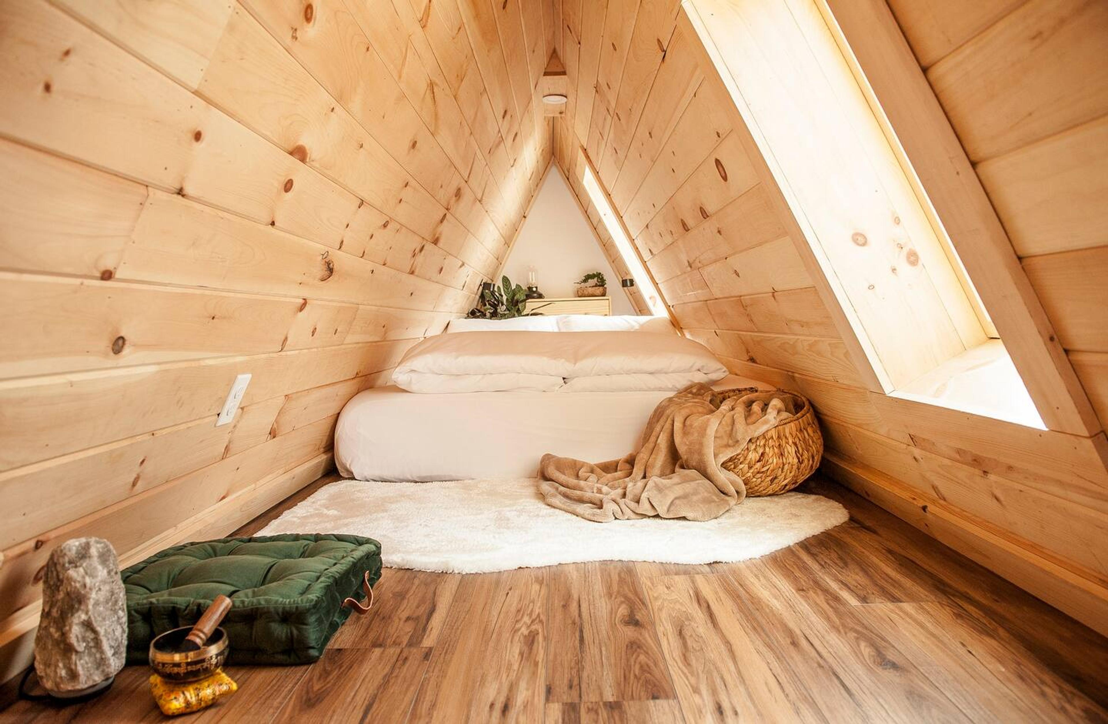A cozy, light-wood A-frame loft features a minimalist sleeping area with white bedding, a plush white rug, and large skylights that brighten the natural timber interior.