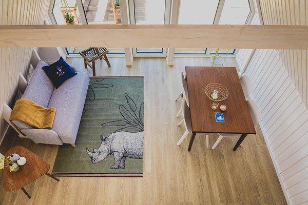 An overhead view of a bright, modern A-frame interior reveals a cozy living area with a rhinoceros-print rug, a gray sofa, and a wooden dining table.