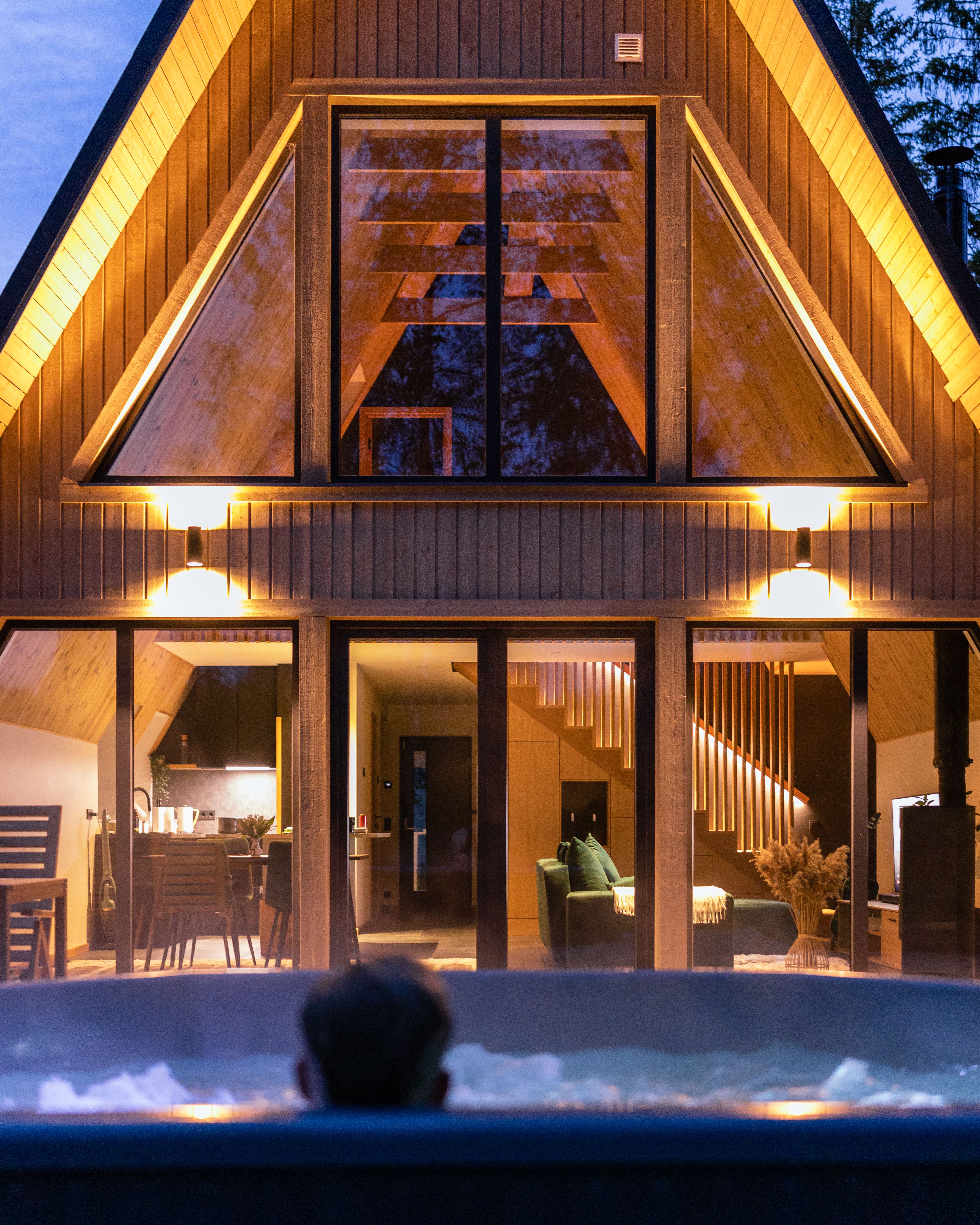 A view from inside a bubbling hot tub looking towards a beautifully illuminated double-height Avrame A-frame home at dusk. The large glass windows reveal a warm, modern interior featuring a cozy living room with a green sofa, a contemporary kitchen, and an illuminated wooden staircase.
