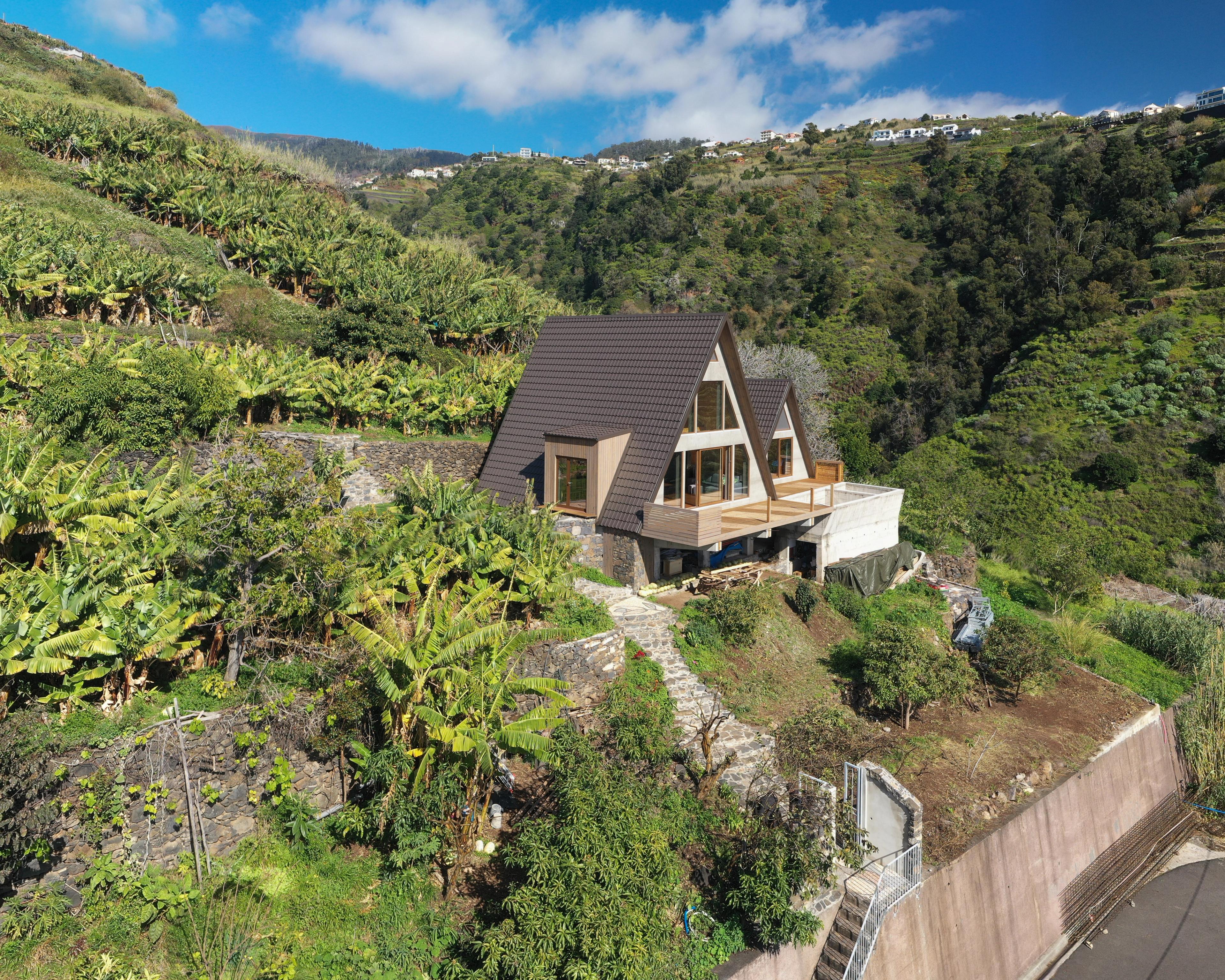 An aerial view of a modern A-frame cabin built into a terraced hillside landscape. The cabin features a dark brown tiled roof, wooden cladding, and a large wooden terrace, surrounded by lush green tropical vegetation and banana trees.