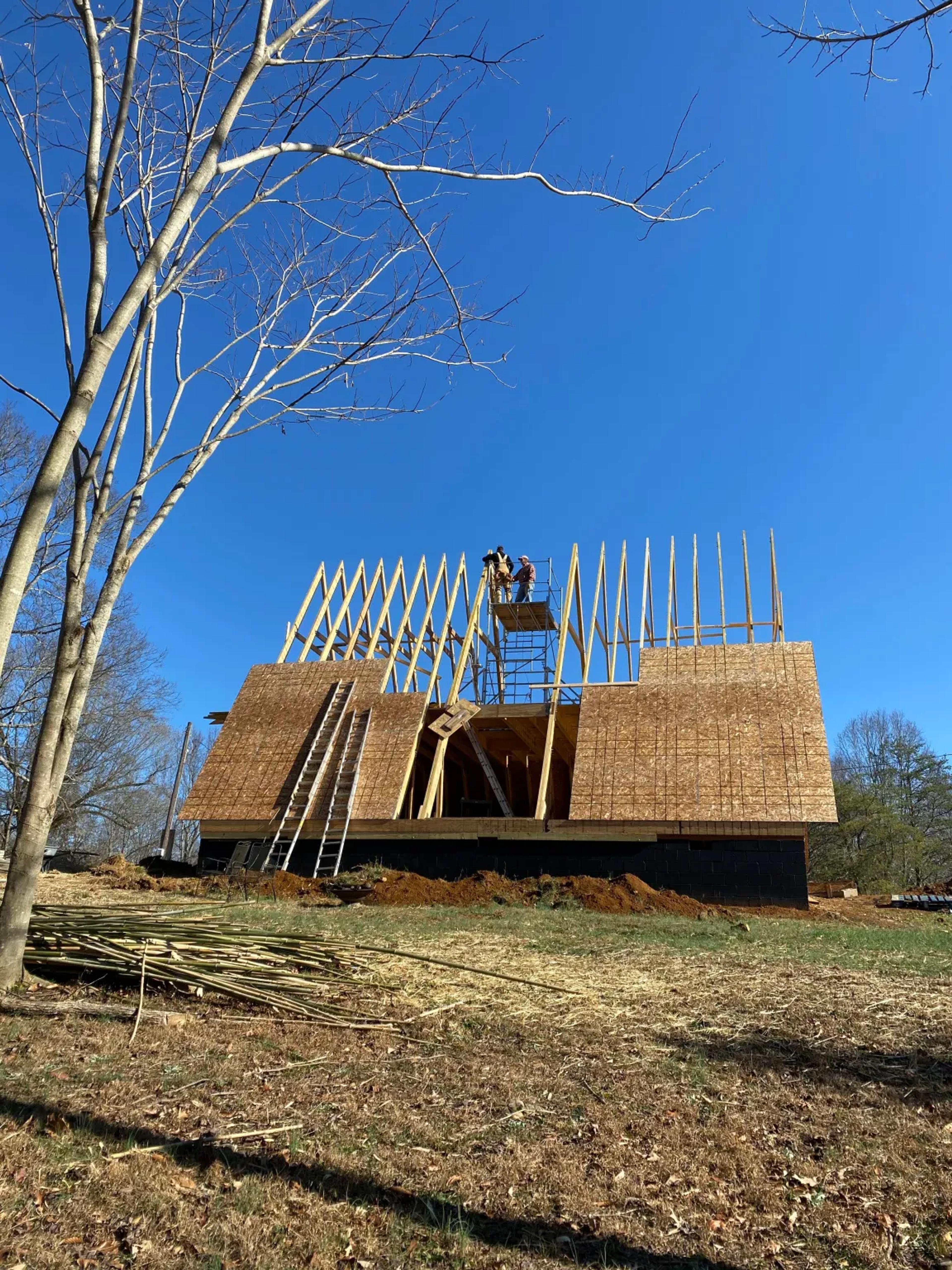 Workers install OSB sheathing onto the lower portion of a large A-frame timber structure while standing on scaffolding under a clear blue sky.