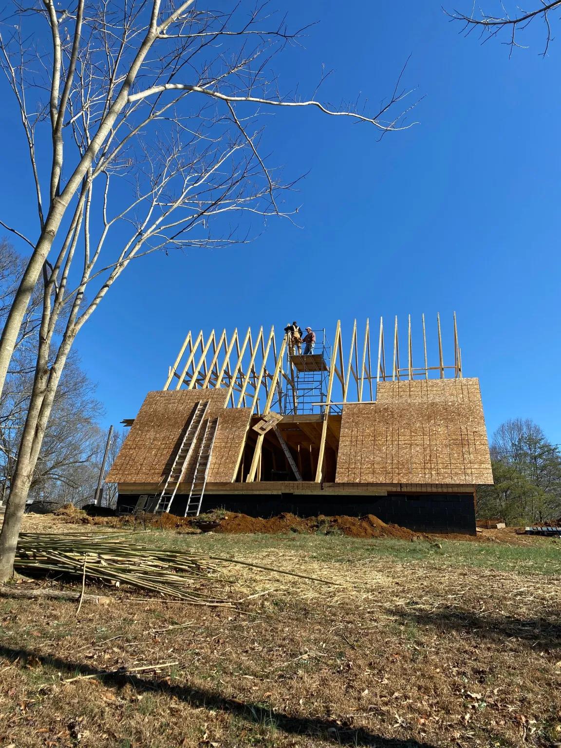 Workers install OSB sheathing onto the lower portion of a large A-frame timber structure while standing on scaffolding under a clear blue sky.