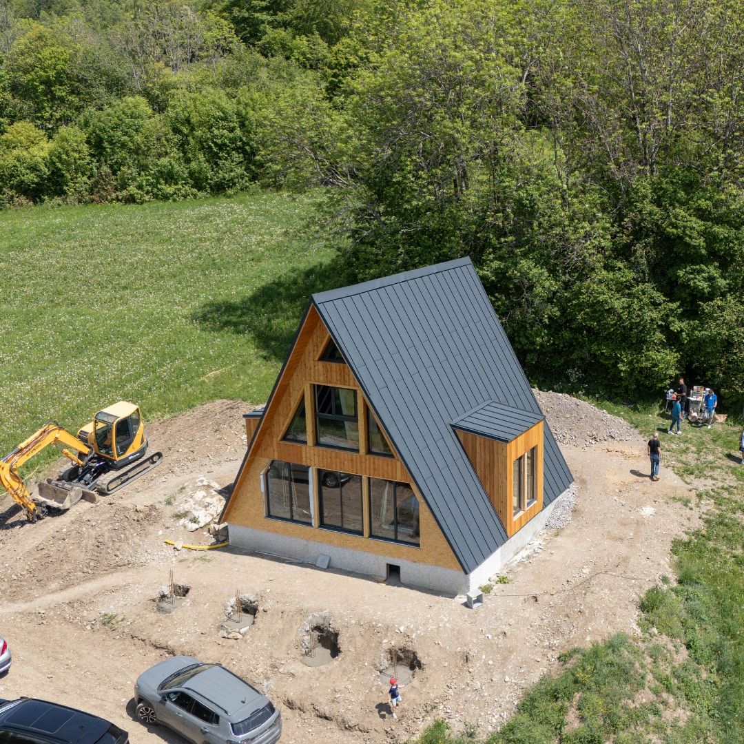 Aerial view of a nearly finished Avrame Trio 75 A-frame house on a hillside in France, with open house day visitors visible to the right