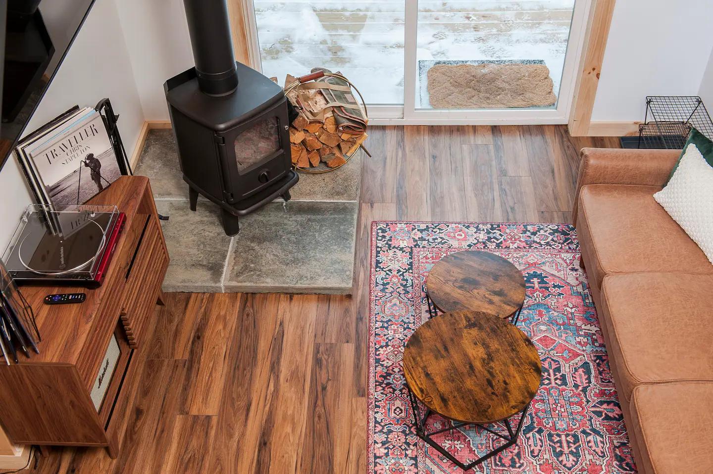 A high-angle view of a cozy living area features a black wood-burning stove, a leather sofa, and a record player setup arranged on warm wood flooring with a colorful patterned rug.