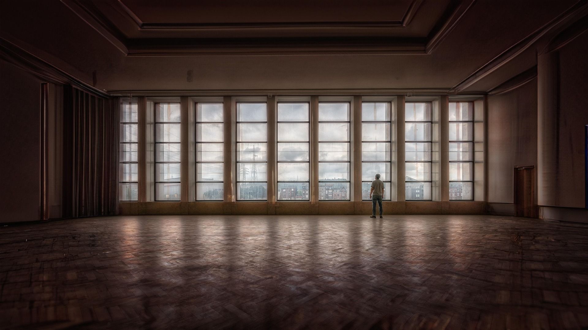 A wide shot of a vast, empty interior hall with a polished wooden parquet floor in a herringbone pattern. A single person stands with their back to the camera, looking out through a massive wall of floor-to-ceiling multi-paned windows. The view outside reveals a cloudy sky, city buildings, and utility towers under a muted light.
