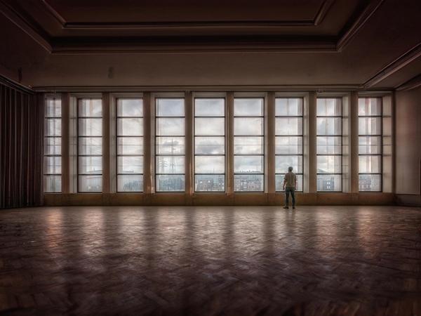 A wide shot of a vast, empty interior hall with a polished wooden parquet floor in a herringbone pattern. A single person stands with their back to the camera, looking out through a massive wall of floor-to-ceiling multi-paned windows. The view outside reveals a cloudy sky, city buildings, and utility towers under a muted light.