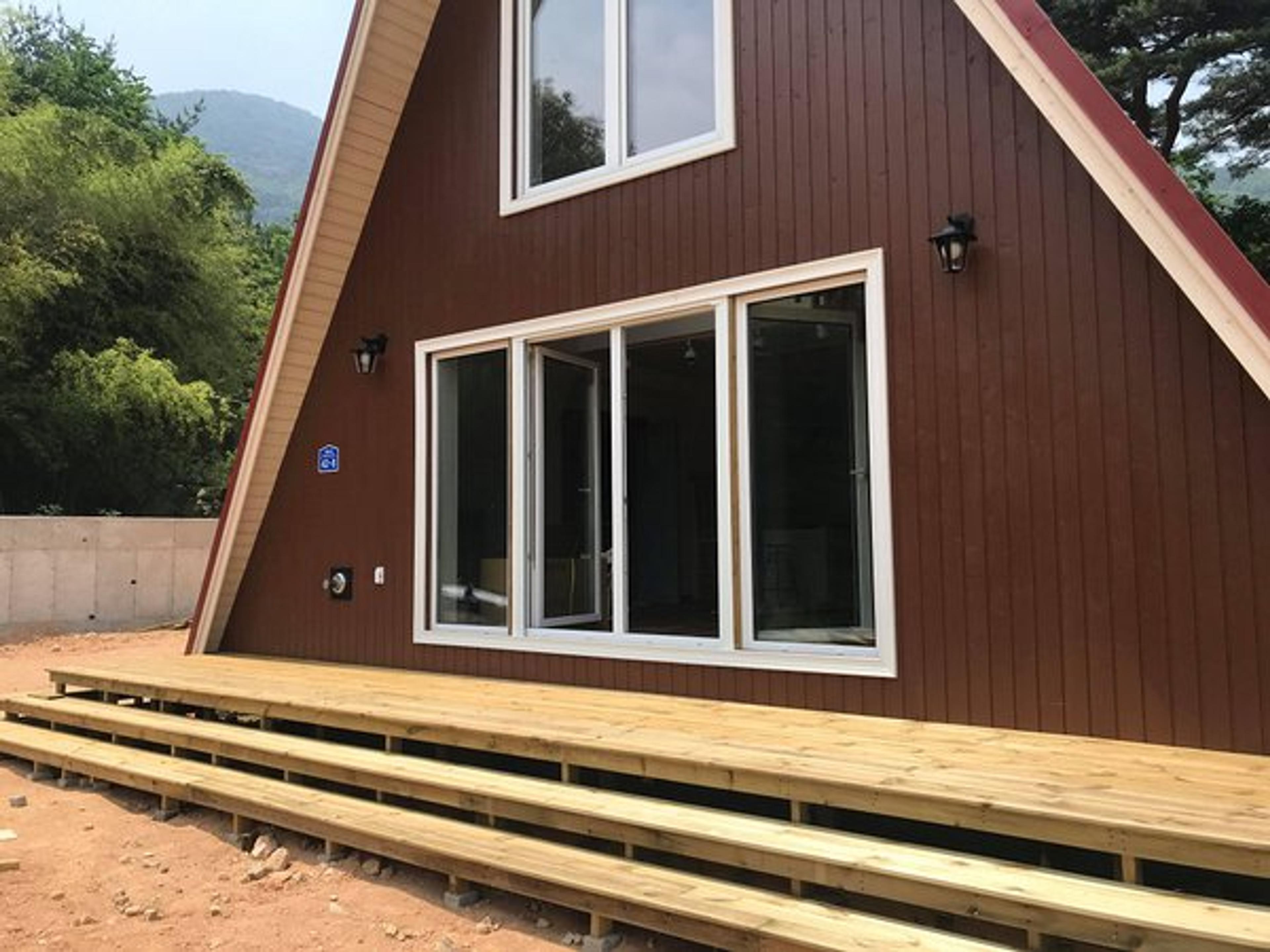 A brown A-frame house features a large wooden deck with multi-level steps, white-trimmed glass doors, and black outdoor sconces.