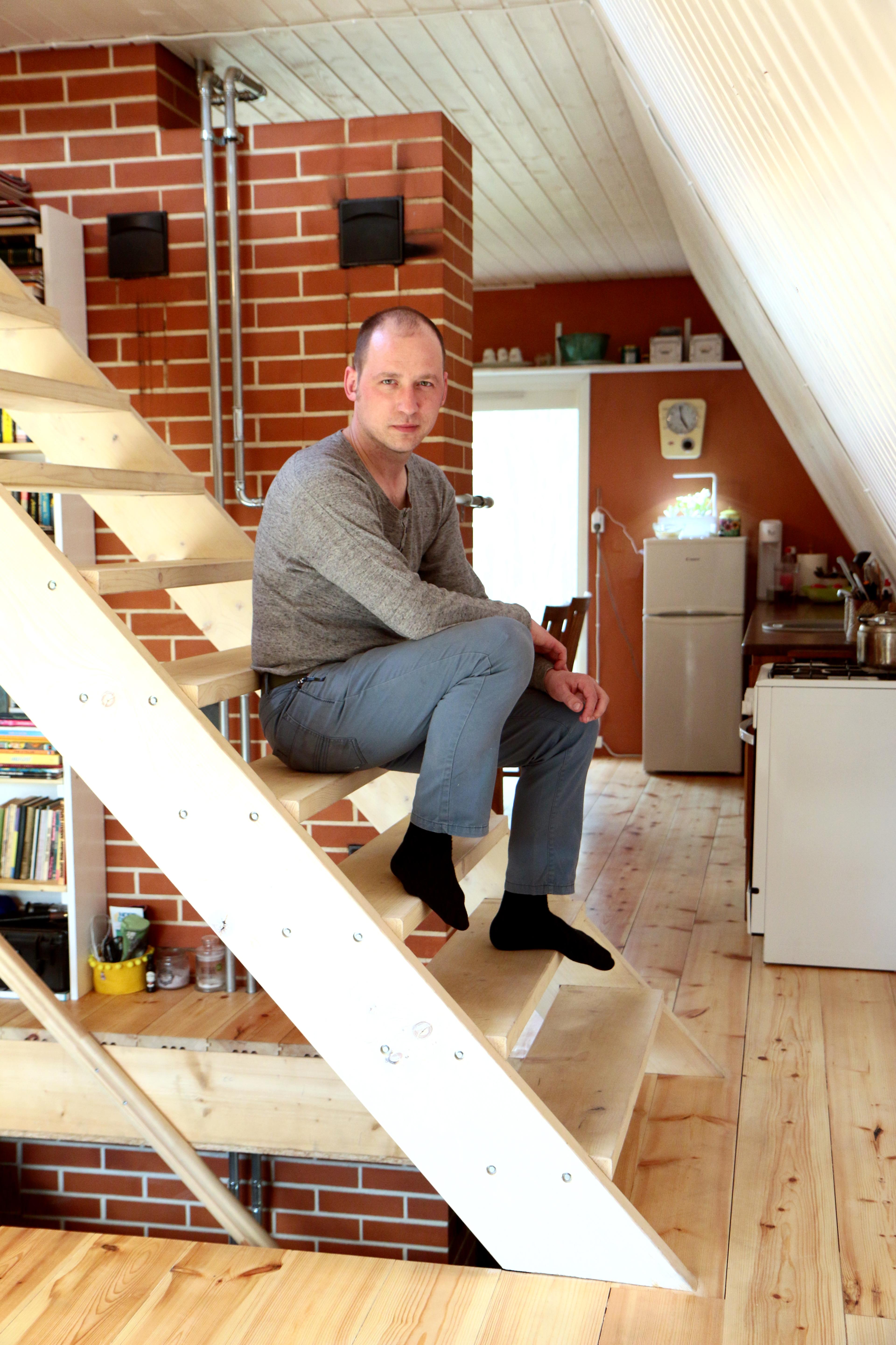 An interior shot of Indrek sitting on light-colored wooden open-tread stairs inside a cozy A-frame house. He is wearing a grey long-sleeve shirt, blue pants, and black socks, looking directly at the camera. The background shows a prominent red brick wall chimney structure, light wood plank flooring, a white slanted ceiling wall, and a small kitchen area equipped with a white stove and compact refrigerator.