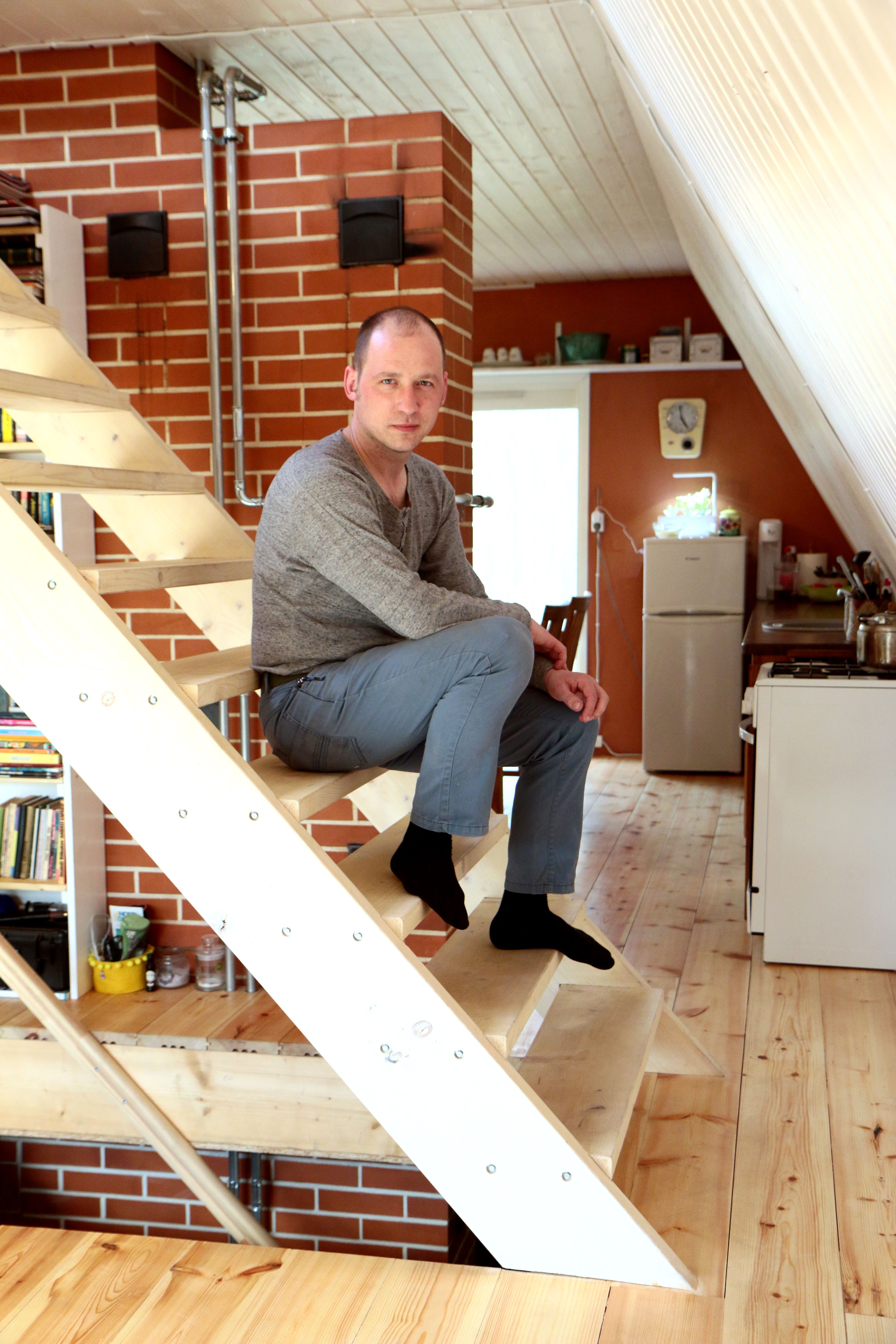 An interior shot of Indrek sitting on light-colored wooden open-tread stairs inside a cozy A-frame house. He is wearing a grey long-sleeve shirt, blue pants, and black socks, looking directly at the camera. The background shows a prominent red brick wall chimney structure, light wood plank flooring, a white slanted ceiling wall, and a small kitchen area equipped with a white stove and compact refrigerator.