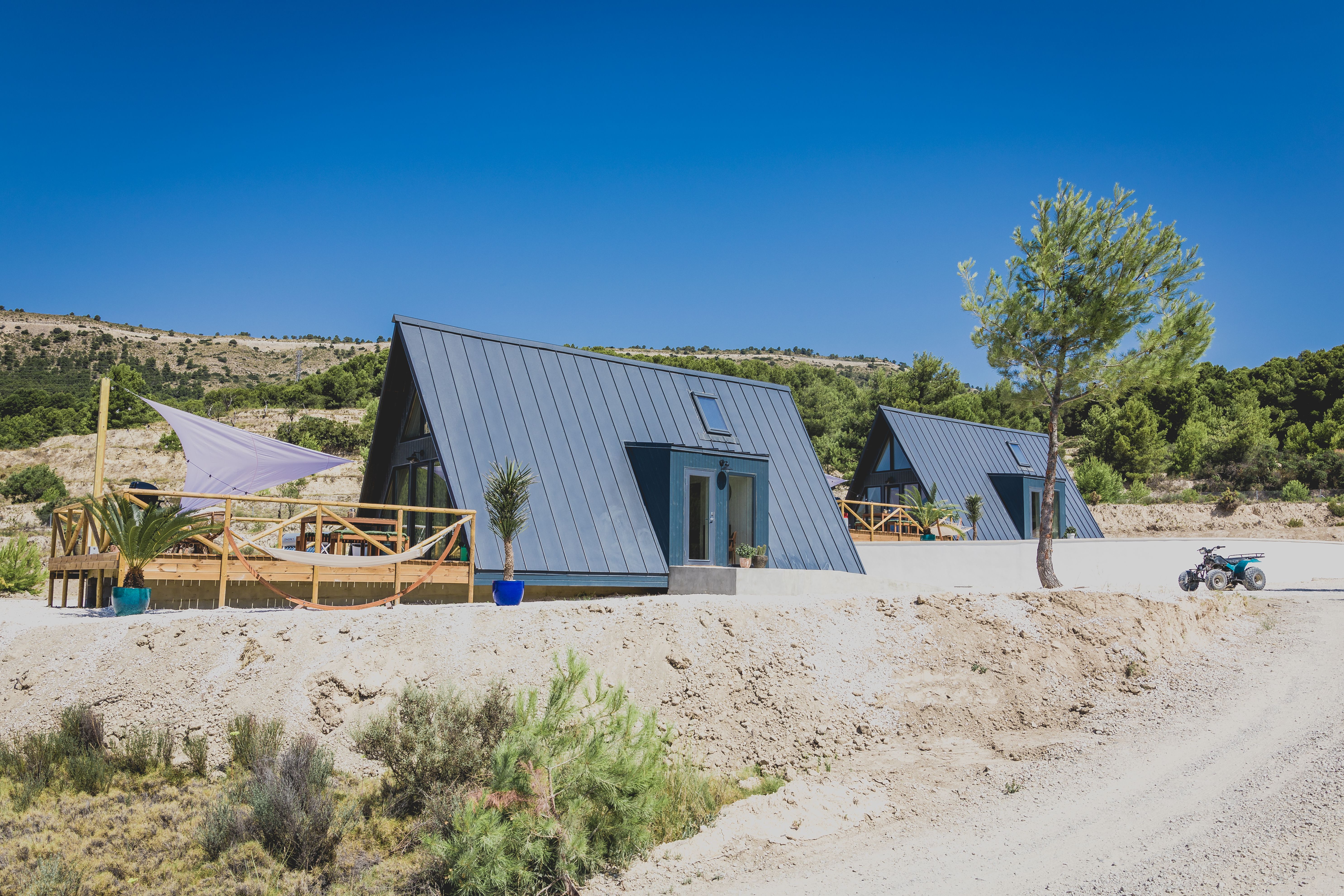 Two modern, dark-grey A-frame cabins with metal roofs sit on a dry, sunny hillside. The foreground cabin features a wooden deck with a white shade sail and a hammock, set against a backdrop of green pine trees and a clear blue sky.