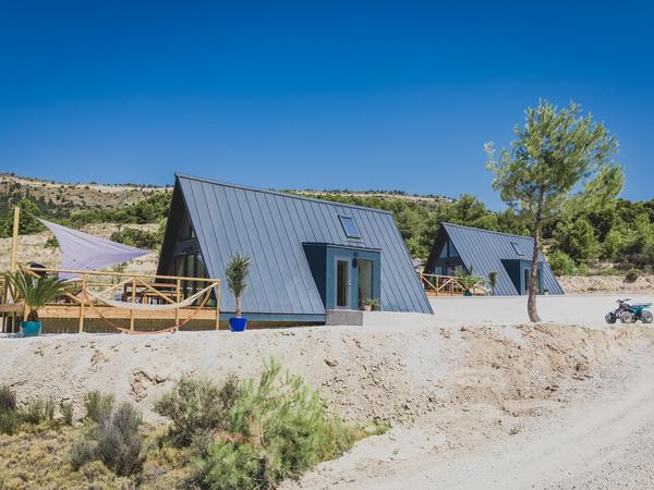Two modern, dark-grey A-frame cabins with metal roofs sit on a dry, sunny hillside. The foreground cabin features a wooden deck with a white shade sail and a hammock, set against a backdrop of green pine trees and a clear blue sky.