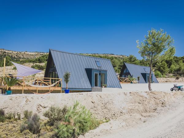 Two modern, dark-grey A-frame cabins with metal roofs sit on a dry, sunny hillside. The foreground cabin features a wooden deck with a white shade sail and a hammock, set against a backdrop of green pine trees and a clear blue sky.
