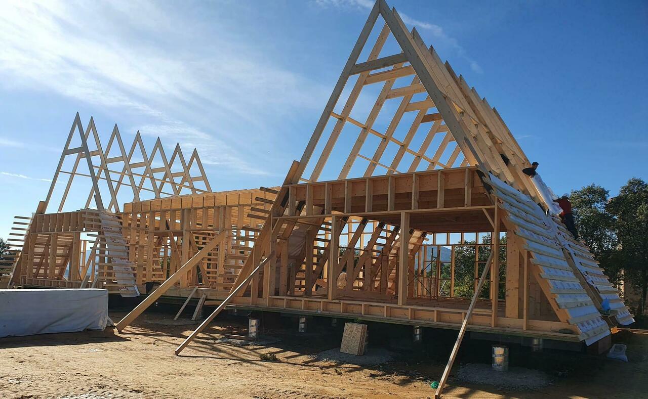 A side-view of a large A-frame house under construction shows the exposed wooden structural framing and rafters against a bright blue sky.