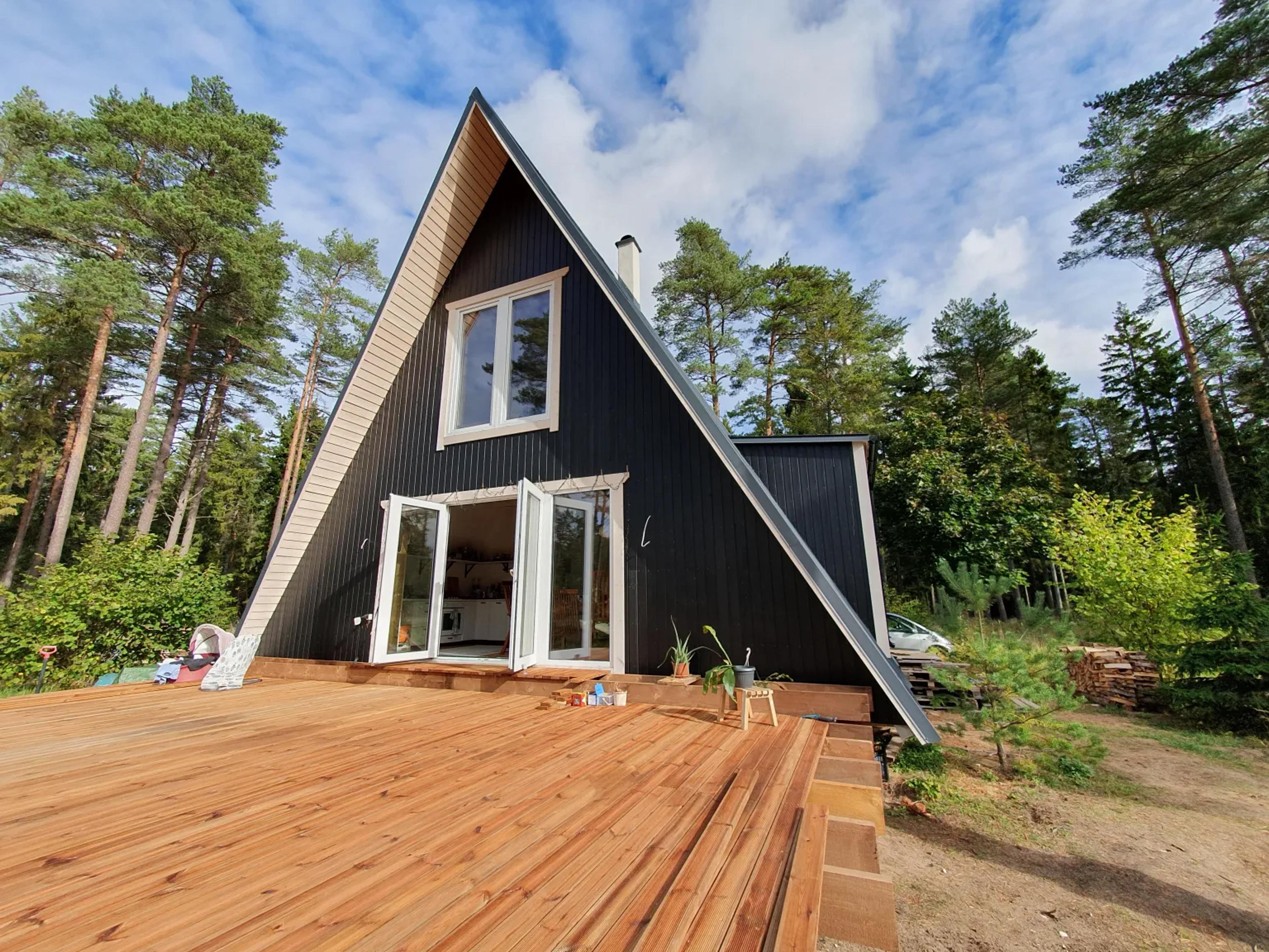A black A-frame cabin with white-trimmed windows and open double doors is featured with an expansive, newly built wooden deck in a forest setting.