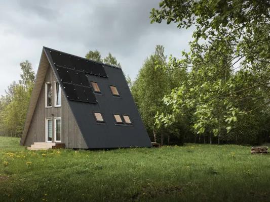 A grey wood-clad A-frame cabin featuring solar panels on its dark metal roof sits in a grassy meadow surrounded by a lush forest under an overcast sky.