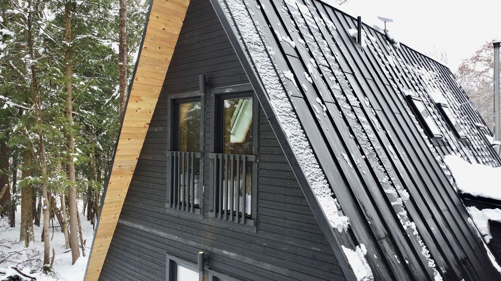 A close-up, high-angle view of the upper story and roof of a modern dark-stained timber A-frame cabin. The structure features a steep black metal standing-seam roof with light snow patches and integrated roof windows, alongside a light natural wood trim along the roofline. Two upper-level glass windows with black safety railings look out toward a snow-covered forest background.