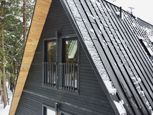 A close-up, high-angle view of the upper story and roof of a modern dark-stained timber A-frame cabin. The structure features a steep black metal standing-seam roof with light snow patches and integrated roof windows, alongside a light natural wood trim along the roofline. Two upper-level glass windows with black safety railings look out toward a snow-covered forest background.