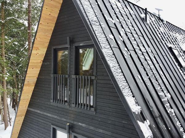 A close-up, high-angle view of the upper story and roof of a modern dark-stained timber A-frame cabin. The structure features a steep black metal standing-seam roof with light snow patches and integrated roof windows, alongside a light natural wood trim along the roofline. Two upper-level glass windows with black safety railings look out toward a snow-covered forest background.