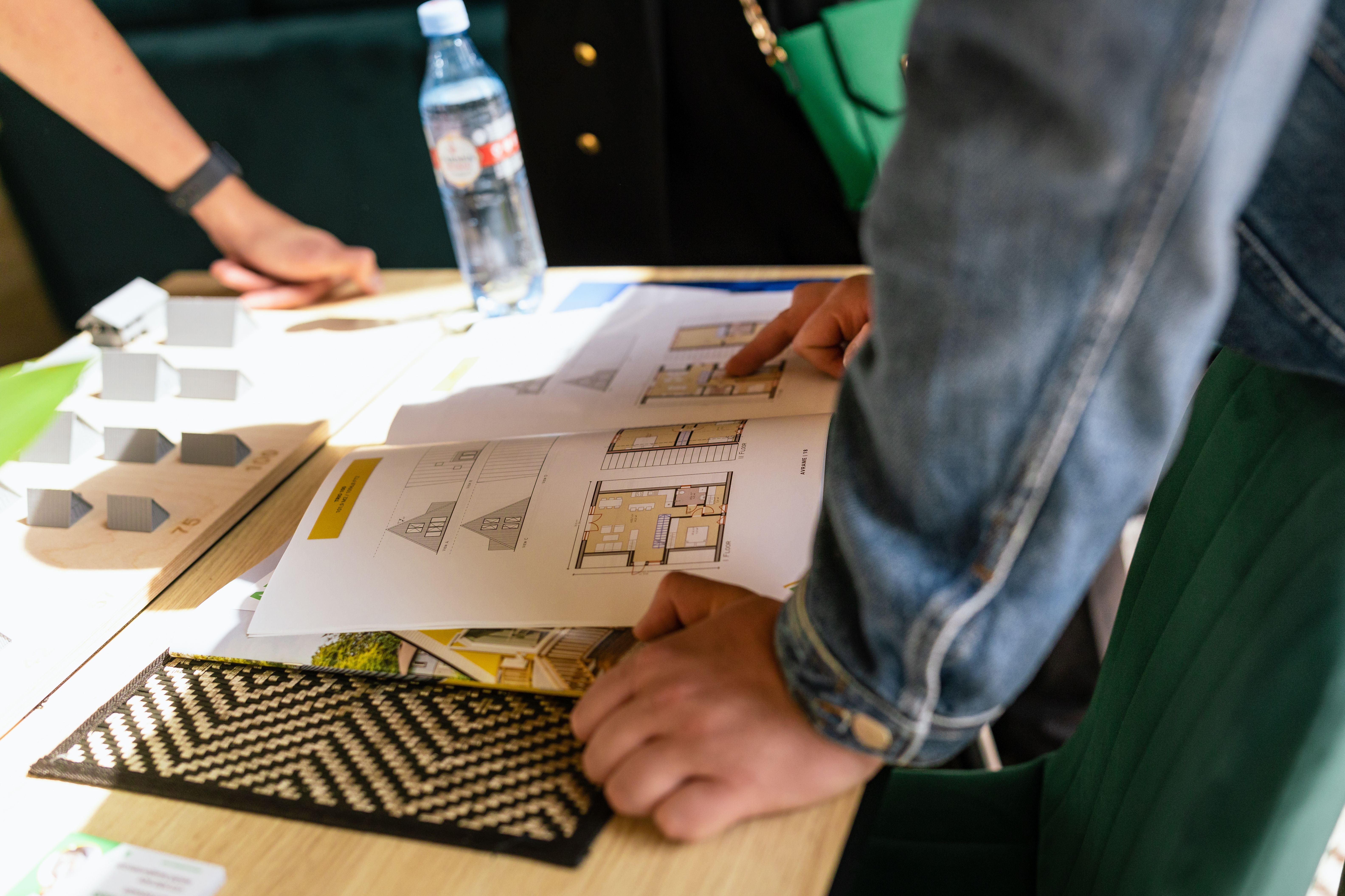 A close-up, high-angle shot of a person in a denim jacket reviewing architectural floor plans and house layouts in a brochure on a wooden table. On the left side of the table, small grey 3D scale models of different A-frame house designs are arranged on a display board.