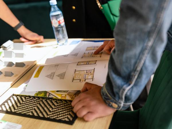 A close-up, high-angle shot of people reviewing floor plans and architectural documents spread across a wooden table. One person in a denim jacket rests their hands near a catalog page showing technical drawings and layouts of an A-frame house model. To the left, several small grey 3D architectural scale models of different A-frame designs sit arranged on a wooden board.