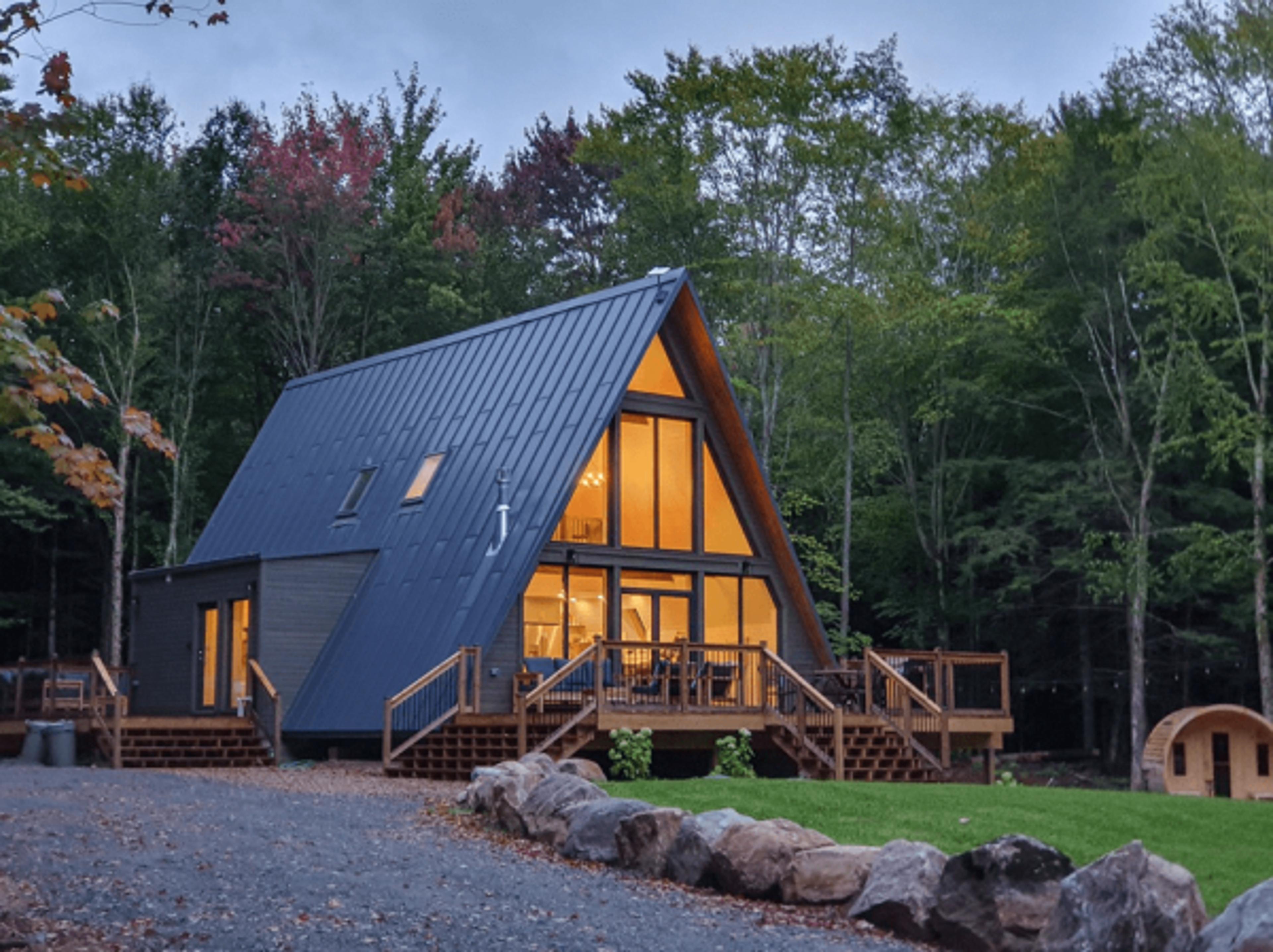 A large, warmly lit A-frame house with a sprawling wooden deck and dark metal roofing sits nestled in a forest clearing at dusk.