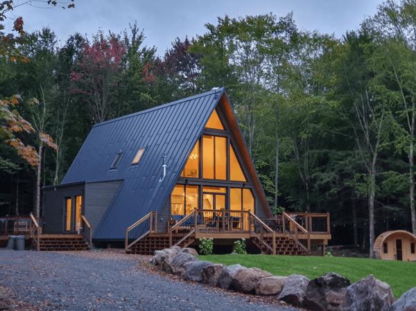 A large, warmly lit A-frame house with a sprawling wooden deck and dark metal roofing sits nestled in a forest clearing at dusk.