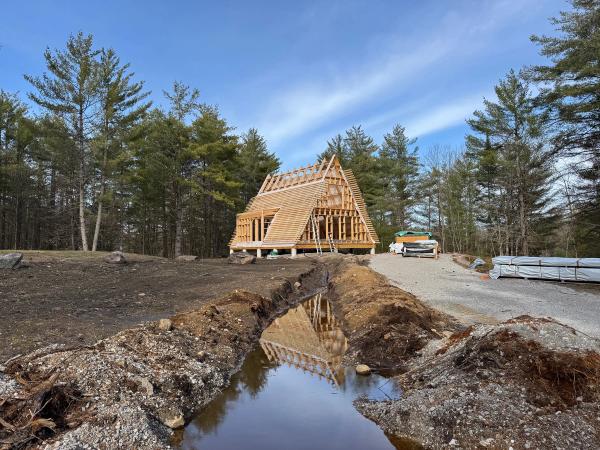 A wide outdoor shot of an Avrame A-frame cabin under construction on a rural lot surrounded by tall pine trees. The raw wooden framing, rafters, and horizontal roof slats are partially assembled against a bright blue sky with wispy clouds. In the foreground, a newly dug drainage trench filled with water perfectly reflects the skeletal structure of the house, while a gravel driveway with wrapped building materials sits to the right.