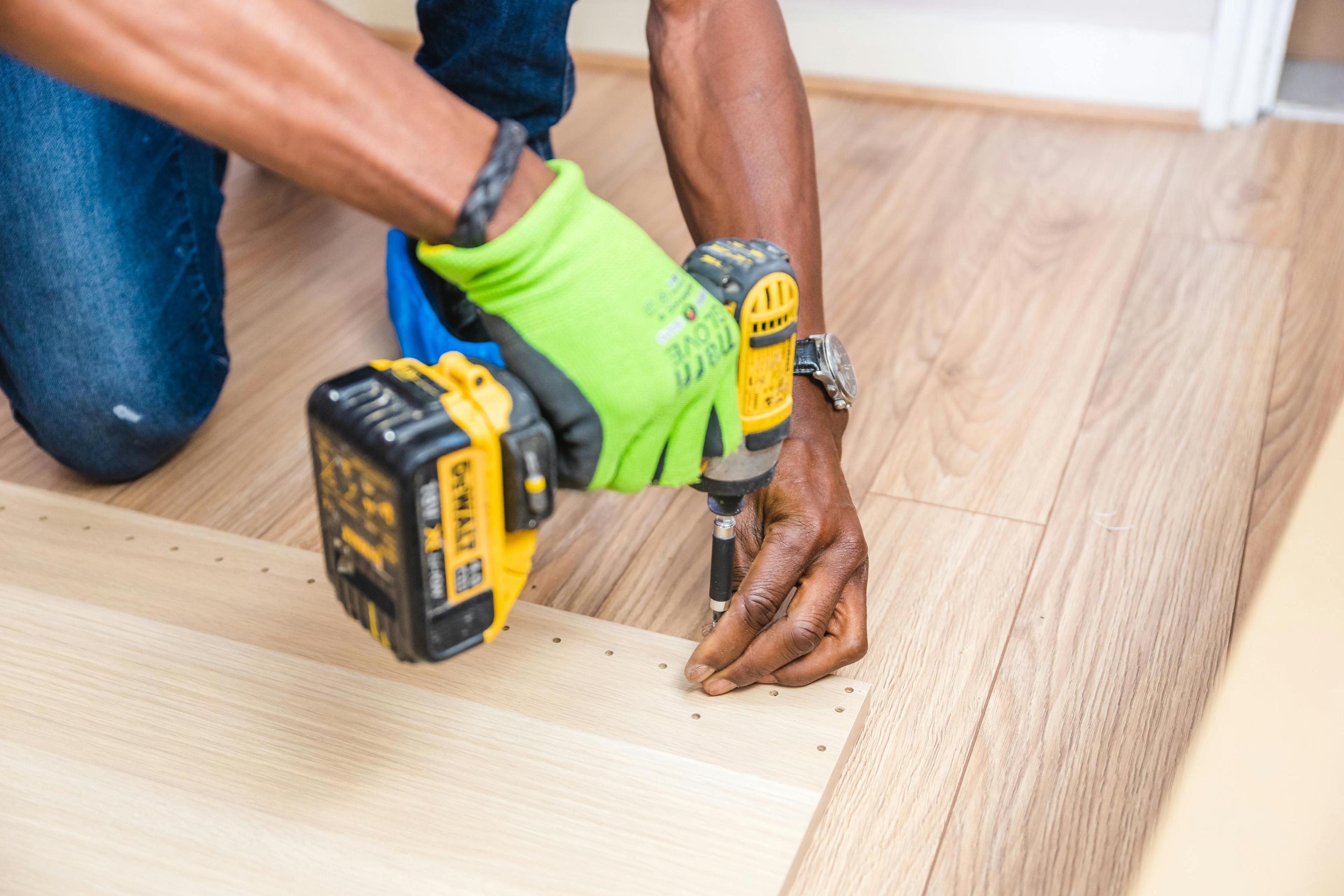 A person wearing a lime green glove uses a yellow power drill to drive a screw into a light-colored wooden board on the floor.