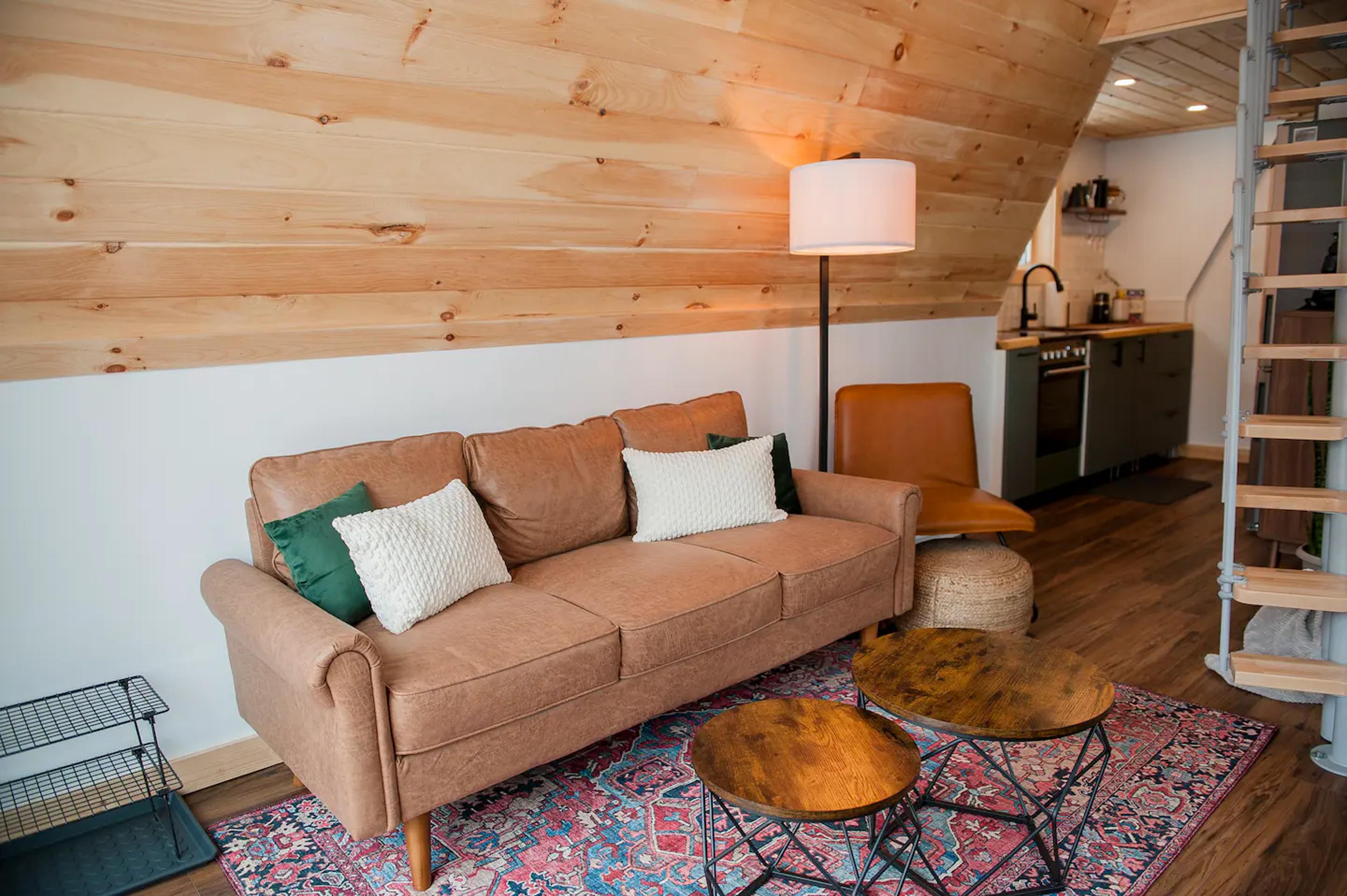 A warm and inviting A-frame living space features a tan leather sofa, rustic round coffee tables, and a colorful rug set against light wood-paneled walls.