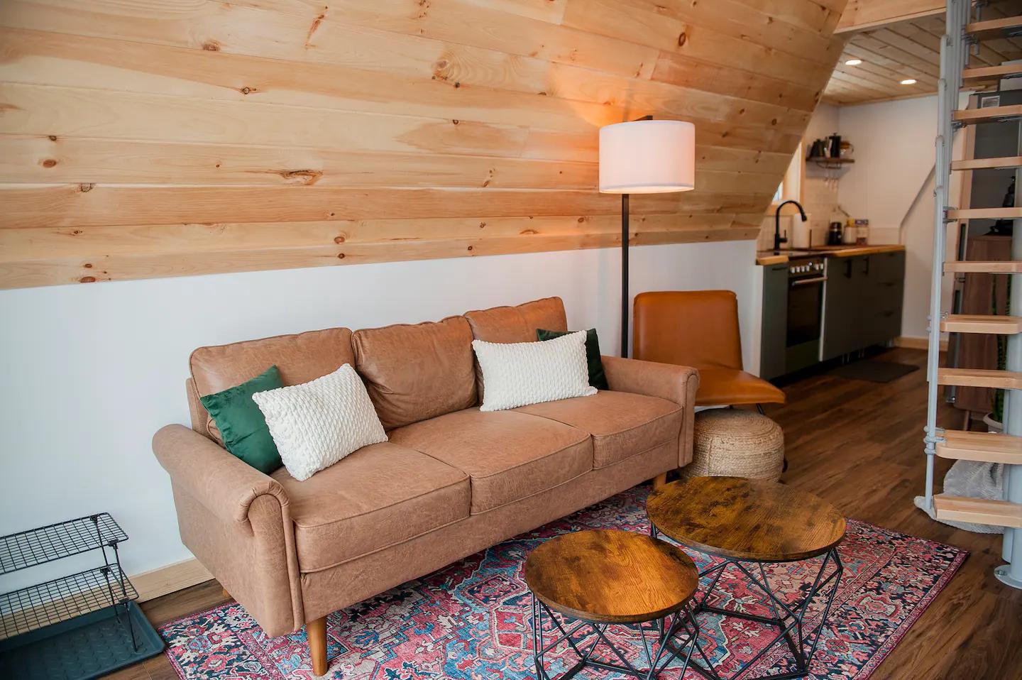 A warm and inviting A-frame living space features a tan leather sofa, rustic round coffee tables, and a colorful rug set against light wood-paneled walls.
