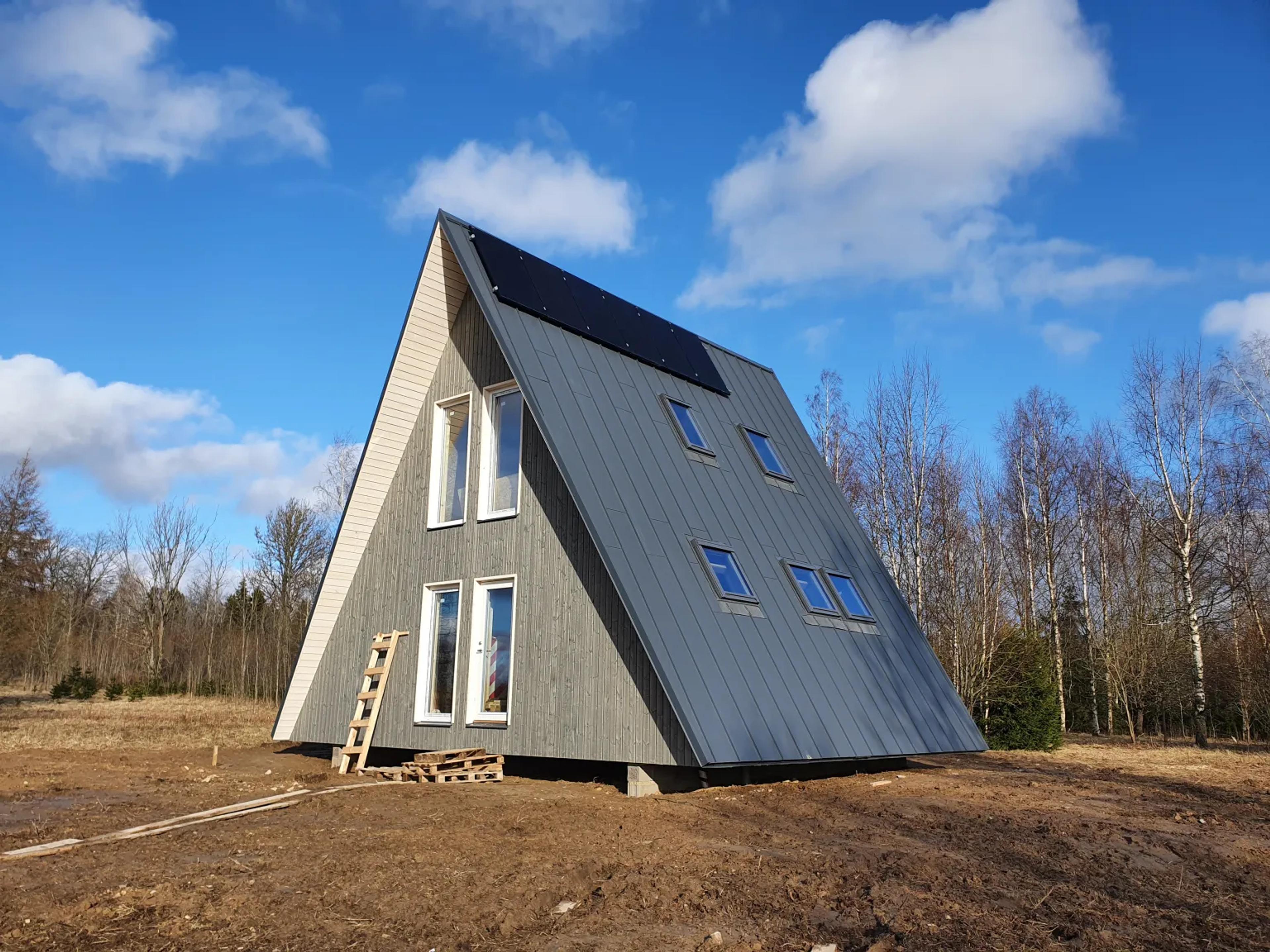 A gray A-frame house under construction features solar panels on its steep metal roof and sits on a dirt plot against a bright blue sky.