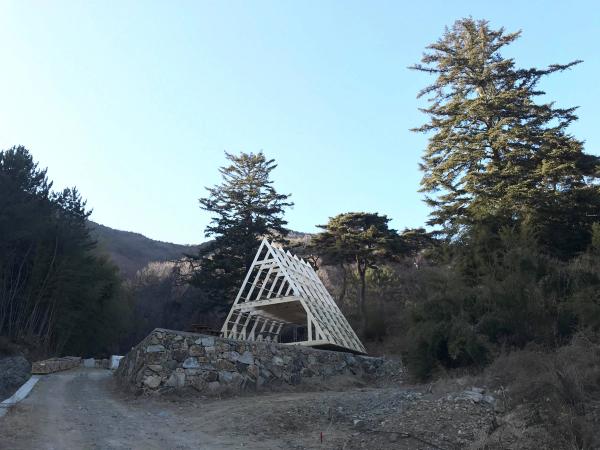 The wooden A-frame skeleton of an Avrame house under construction, perched on a raised stone wall foundation on a mountain lot surrounded by tall pine trees and hills under a clear blue sky.