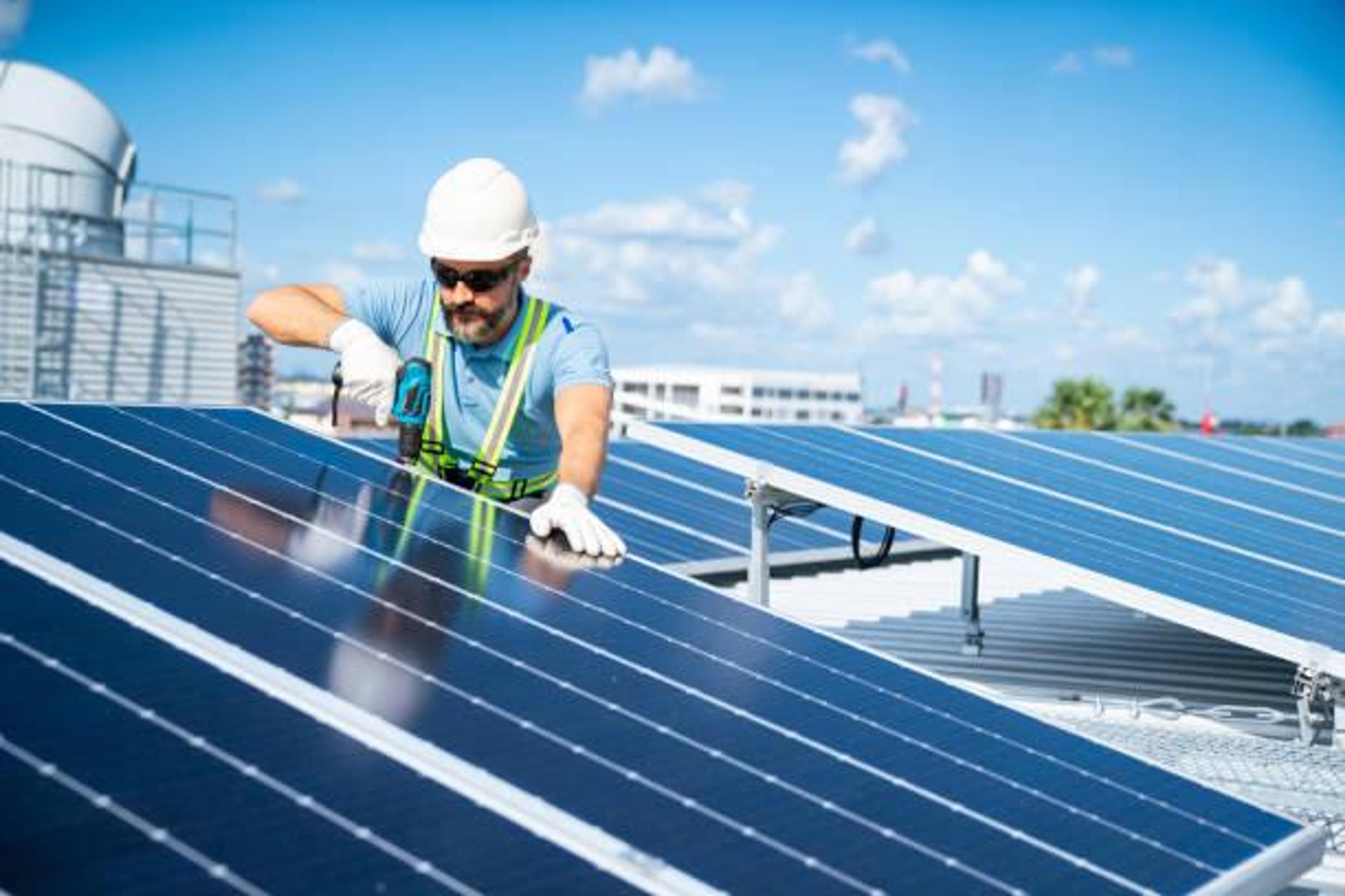 A worker wearing a white hard hat and safety gear uses a power drill to install solar panels on a rooftop under a clear blue sky.