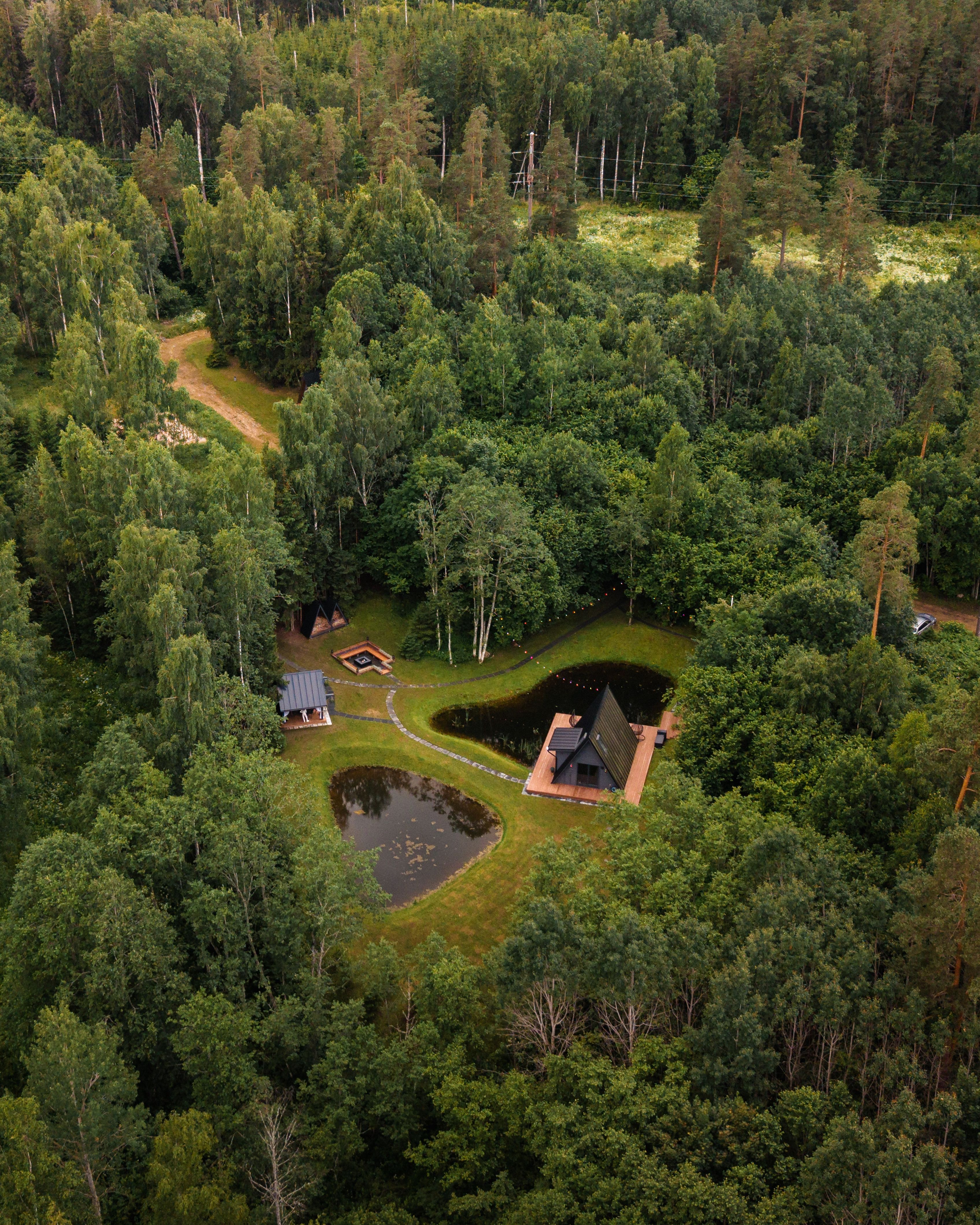 An aerial drone view of a dark A-frame cabin built on a wooden deck next to two small ponds in a dense, green forest. Stone pathways connect the house to a outdoor seating area, a pit fire site, and secondary structures, surrounded by lush, thick treetops.