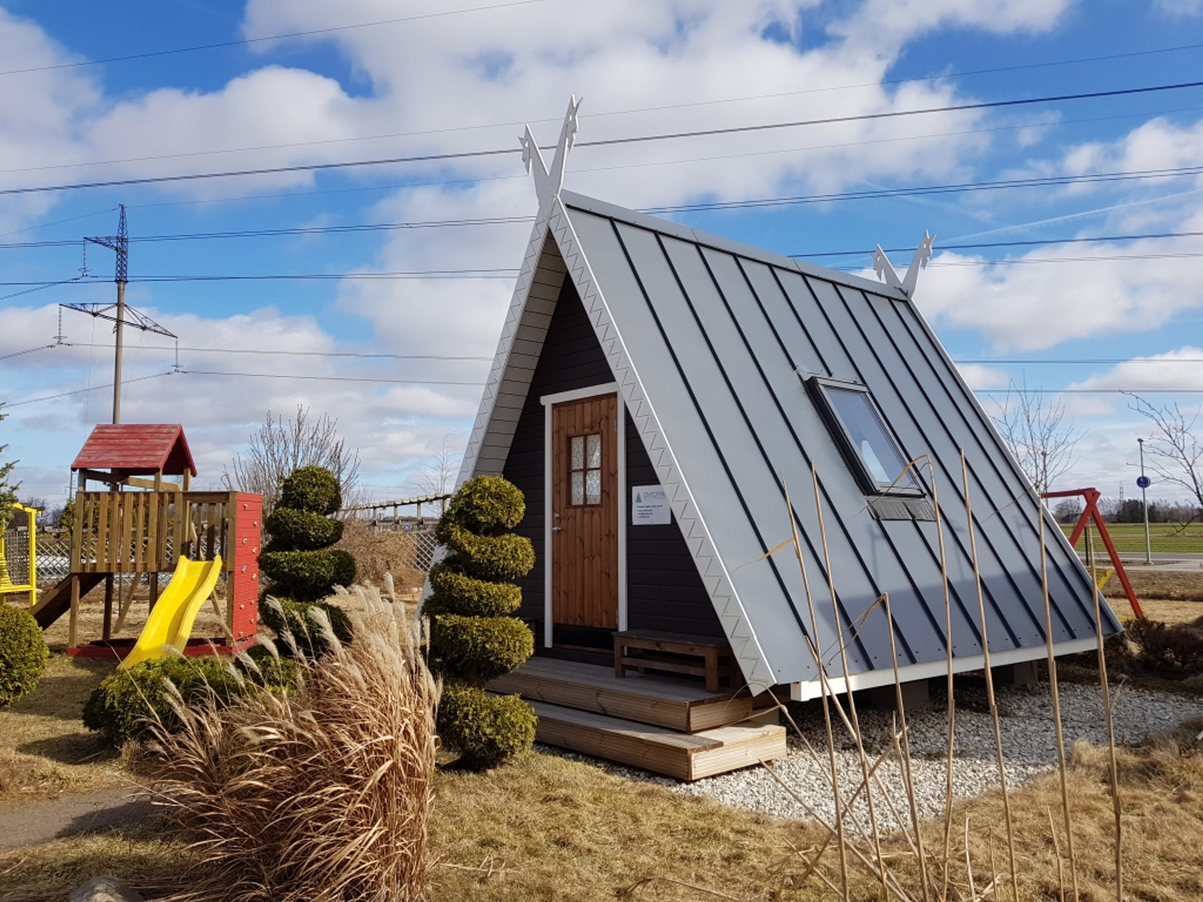 A small A-frame playhouse or studio with decorative roof gables and a skylight sits in a landscaped yard next to a children's slide and spiral-shaped topiaries.