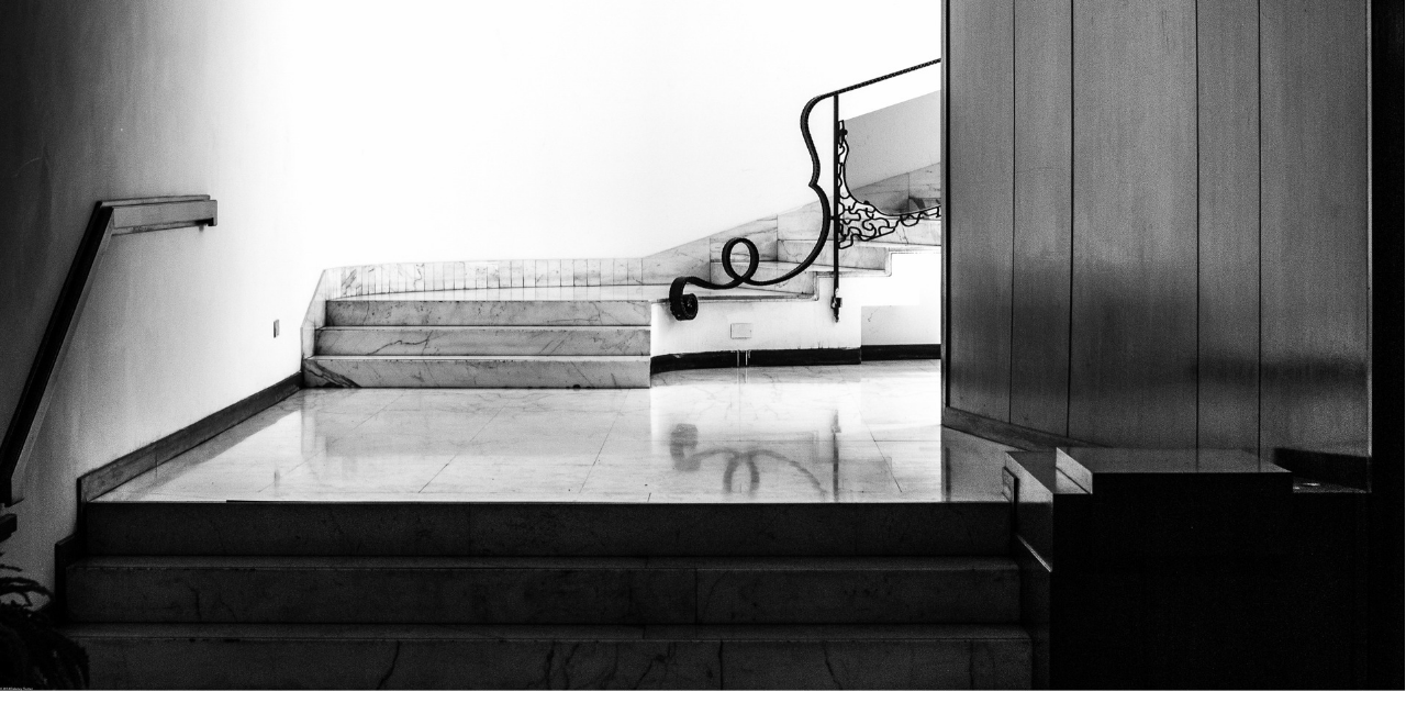 A black-and-white architectural photograph captures a minimalist marble staircase featuring an ornate, swirling wrought-iron handrail against a stark white wall.