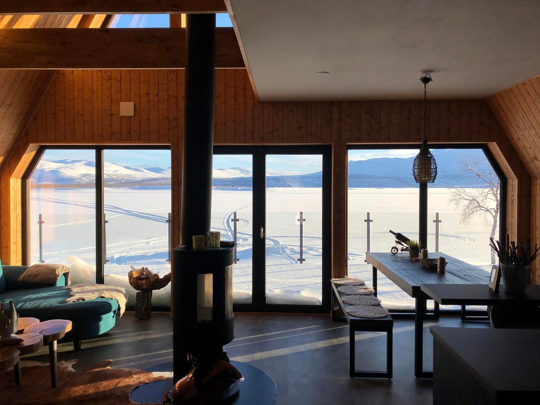 An interior view of a cozy, modern living space inside an A-frame structure, featuring warm, wood-paneled walls and exposed ceiling beams. A central, tall black wood-burning stove separates a sitting area with a blue sofa and natural wood furniture on the left from a dining area with a long wooden table and bench on the right. Both areas look out through a bank of large, floor-to-ceiling windows that perfectly frame a sunlit view of a vast, snowy outdoor expanse and distant mountains. A patterned pendant lamp hangs above the dining table, and animal hide rugs add texture to the floor.