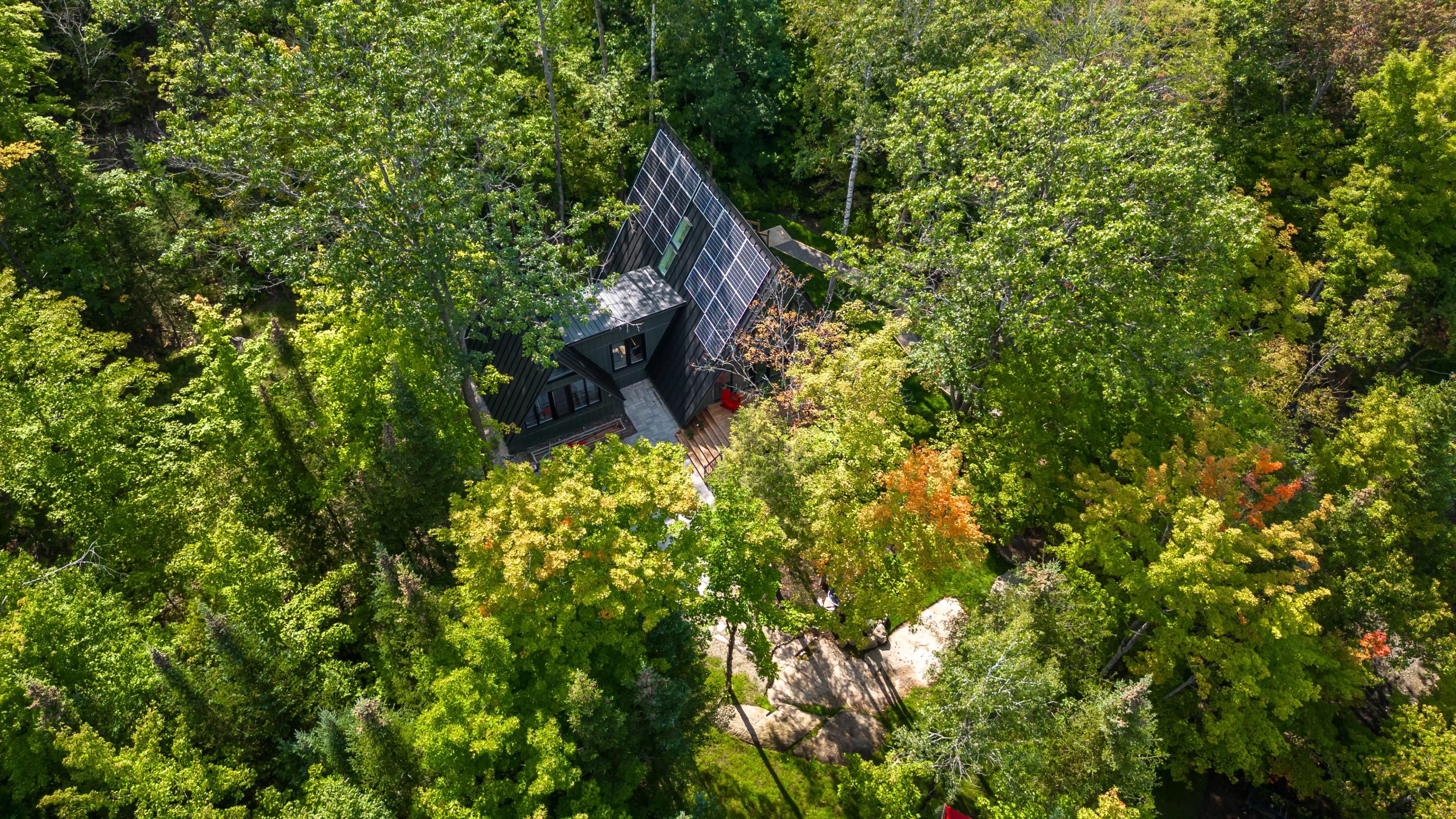 An aerial view shows a dark A-frame house with a large solar panel array on its roof, deeply secluded within a dense green forest.