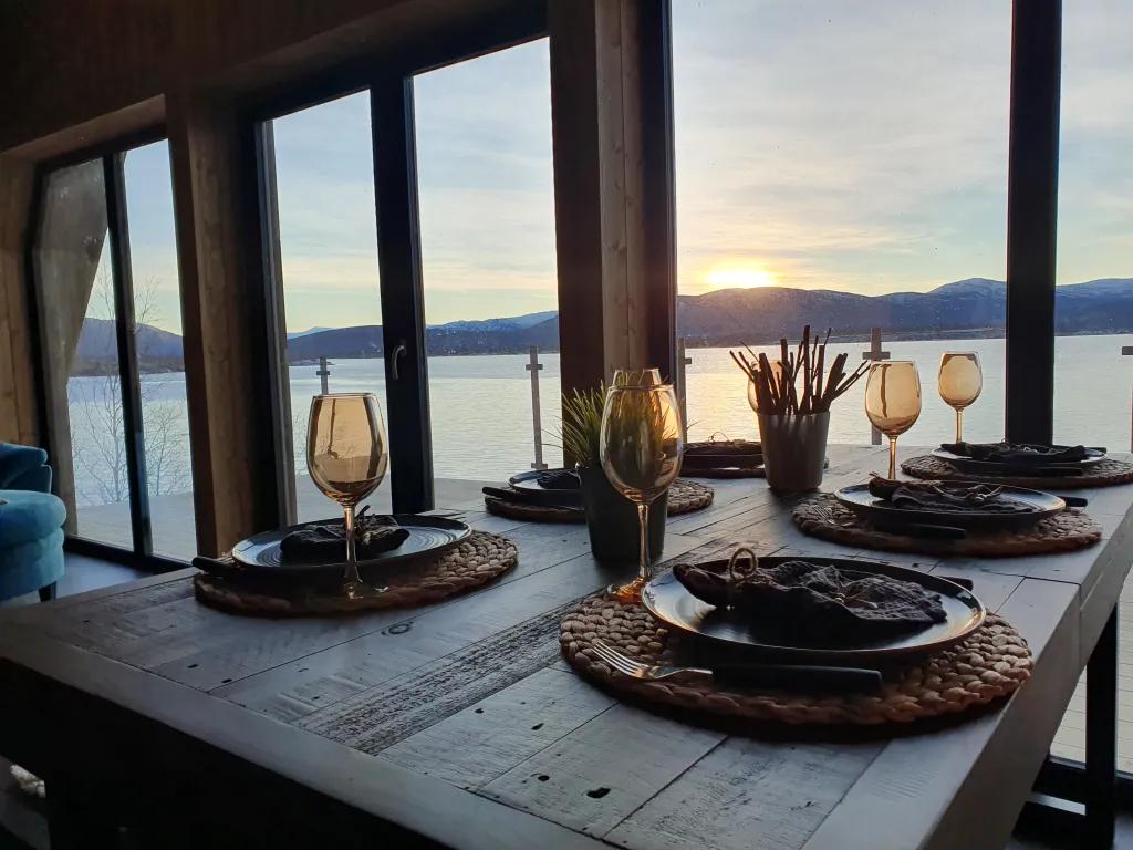 A rustic wooden dining table is elegantly set for a meal with woven placemats and wine glasses, positioned in front of a large window offering a stunning view of a mountain-rimmed lake at sunset.