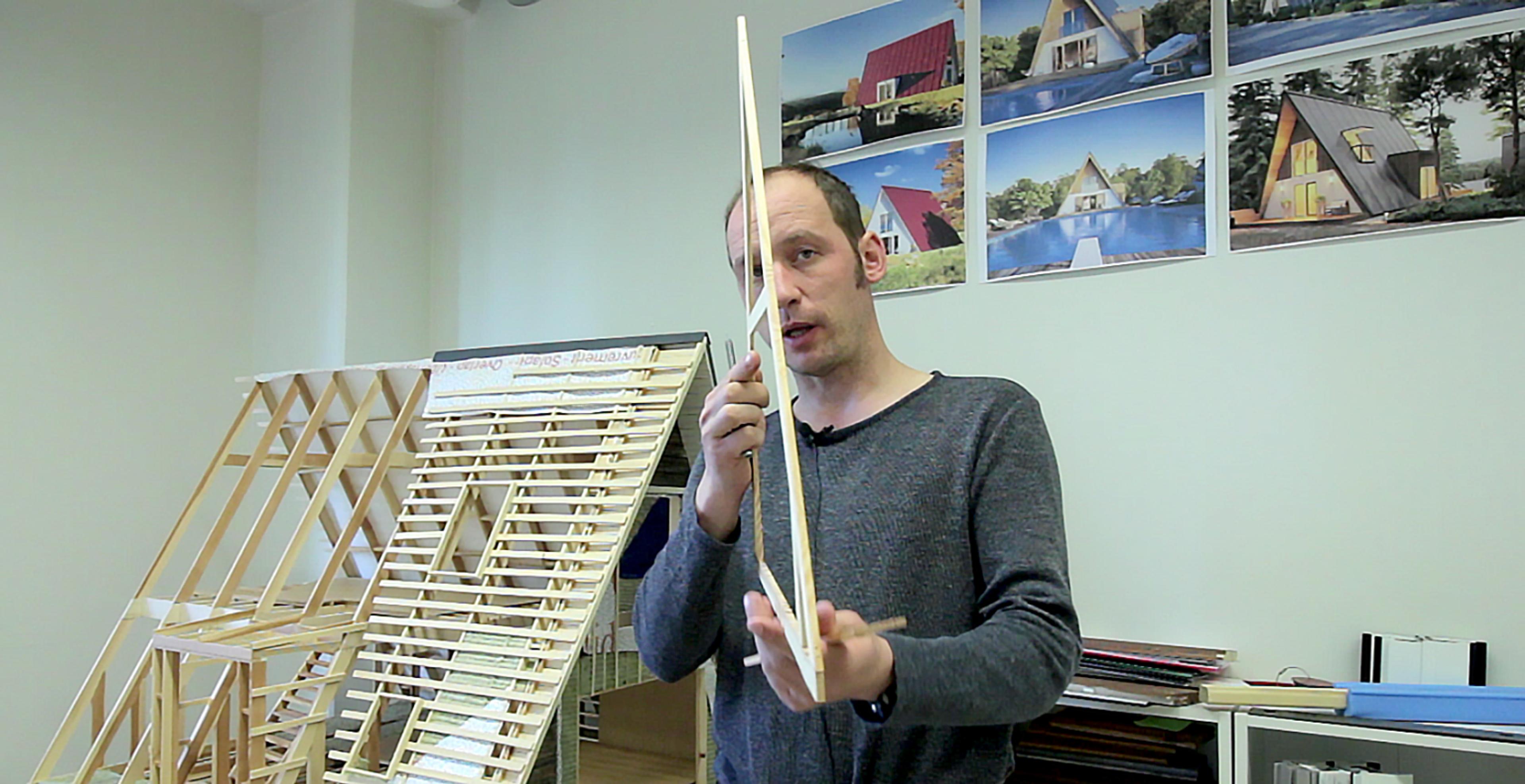 An architectural designer holds a miniature scale model of a triangular roof truss, illustrating the structural "DNA" of the Avrame building system.