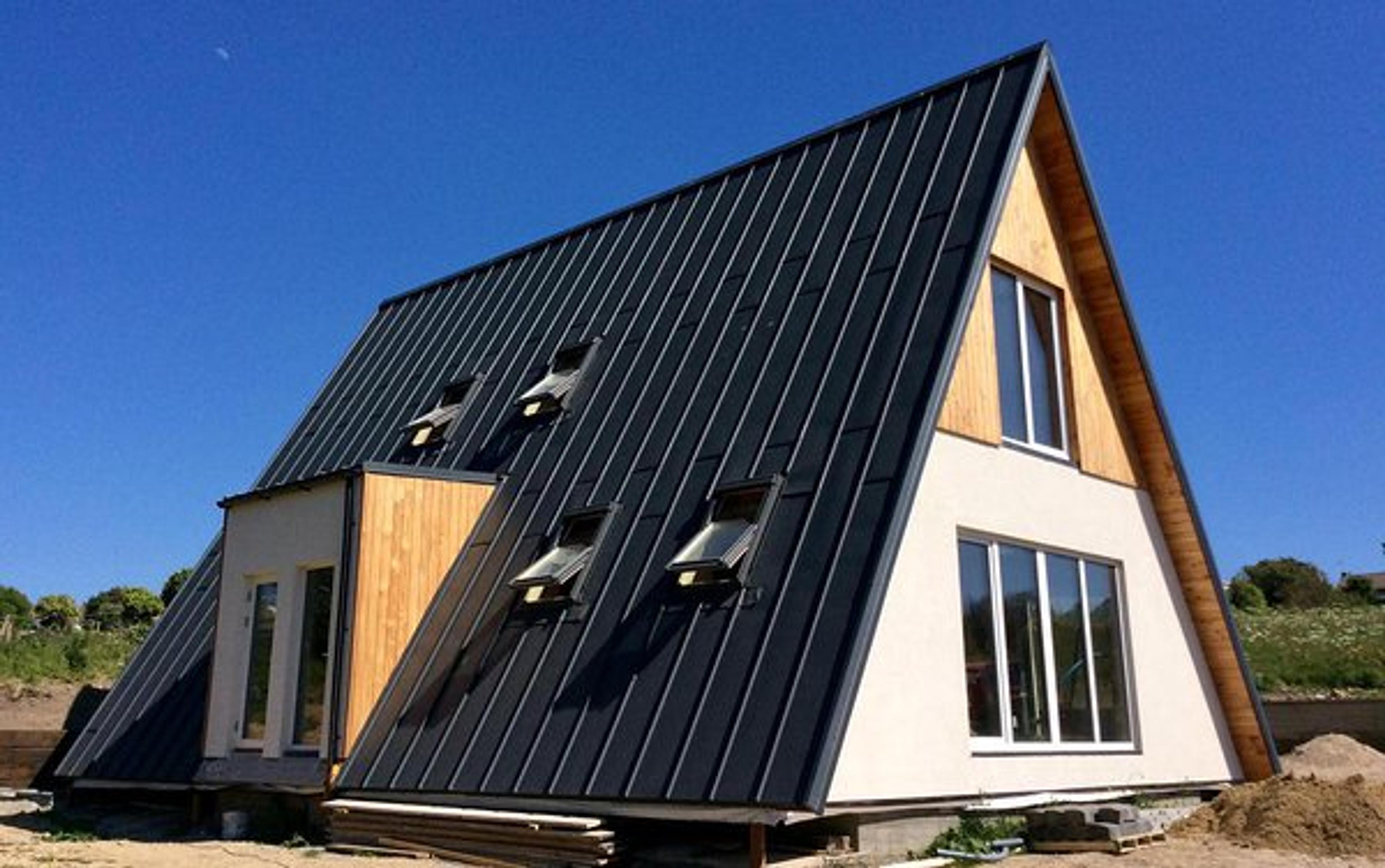 A modern A-frame house features a steep black metal roof with multiple skylights and a combination of white stucco and natural wood exterior siding.