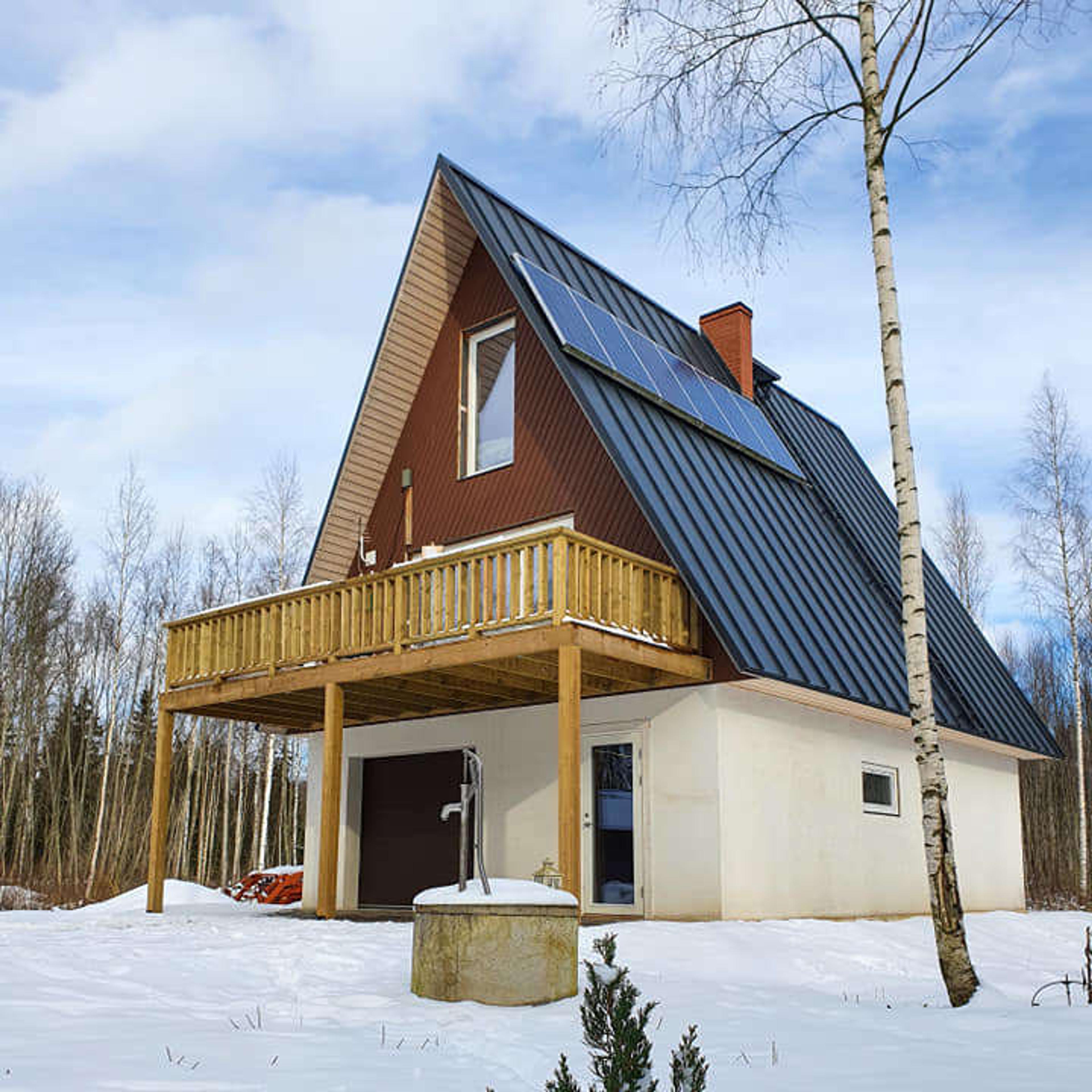 An A-frame house with solar panels and a wooden balcony sits in a snowy landscape, featuring a white ground floor and a brown upper exterior.