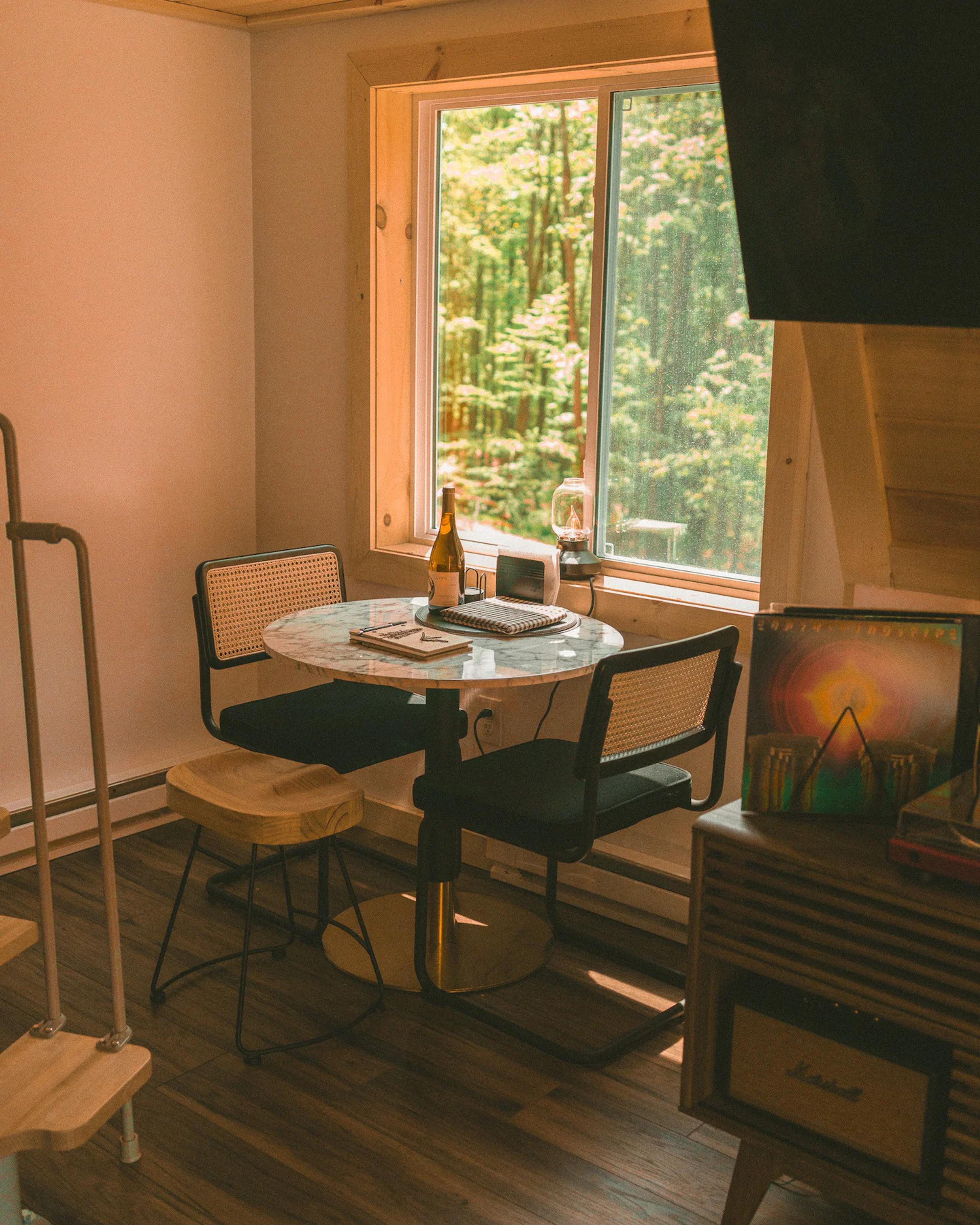 A cozy dining nook features a marble-topped bistro table and cane chairs set beside a large window with views of a lush green forest.