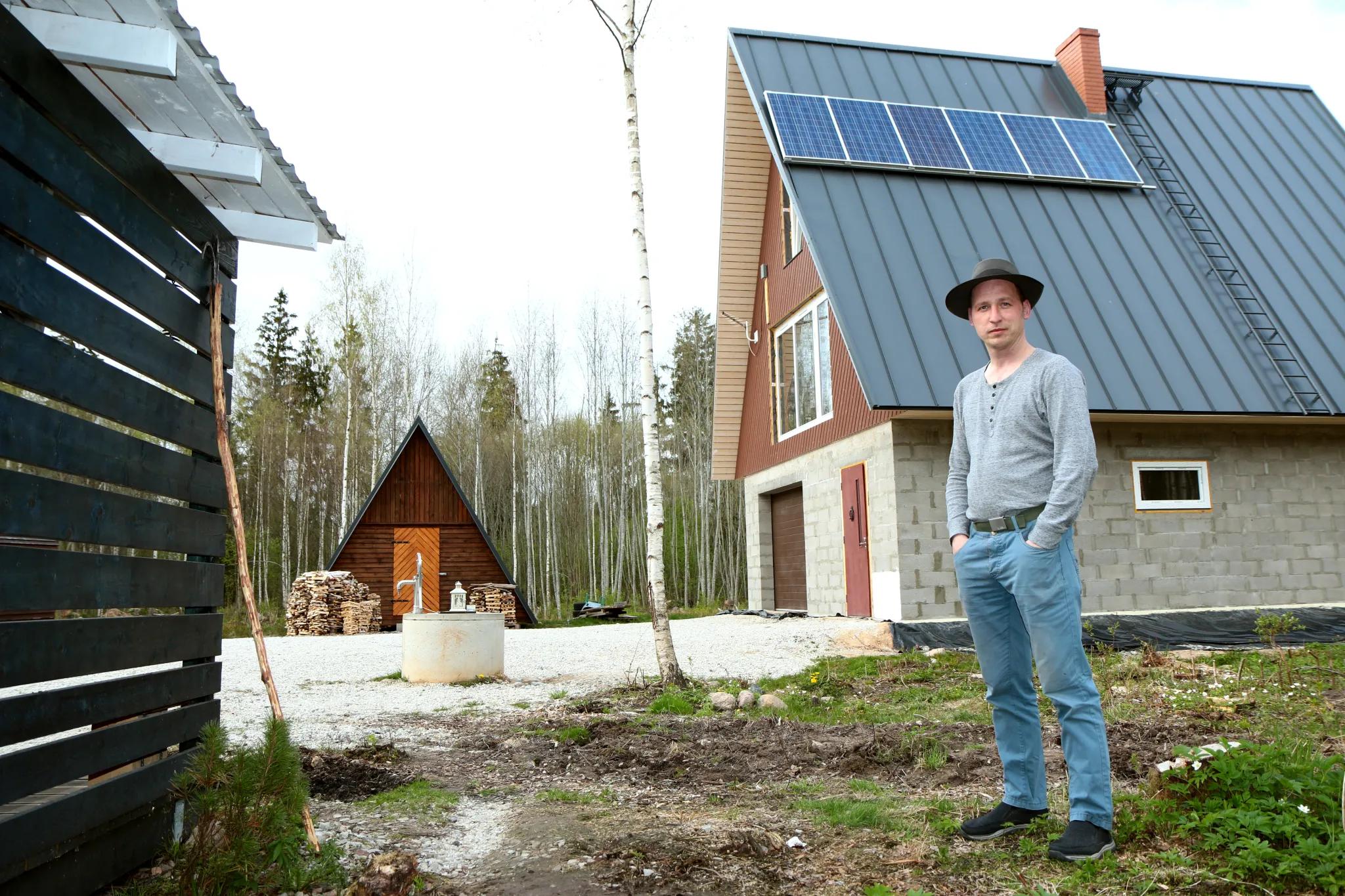 A man in a hat stands in a rural yard before a house with solar panels on its metal roof, with a smaller wooden A-frame cabin and a well in the background.